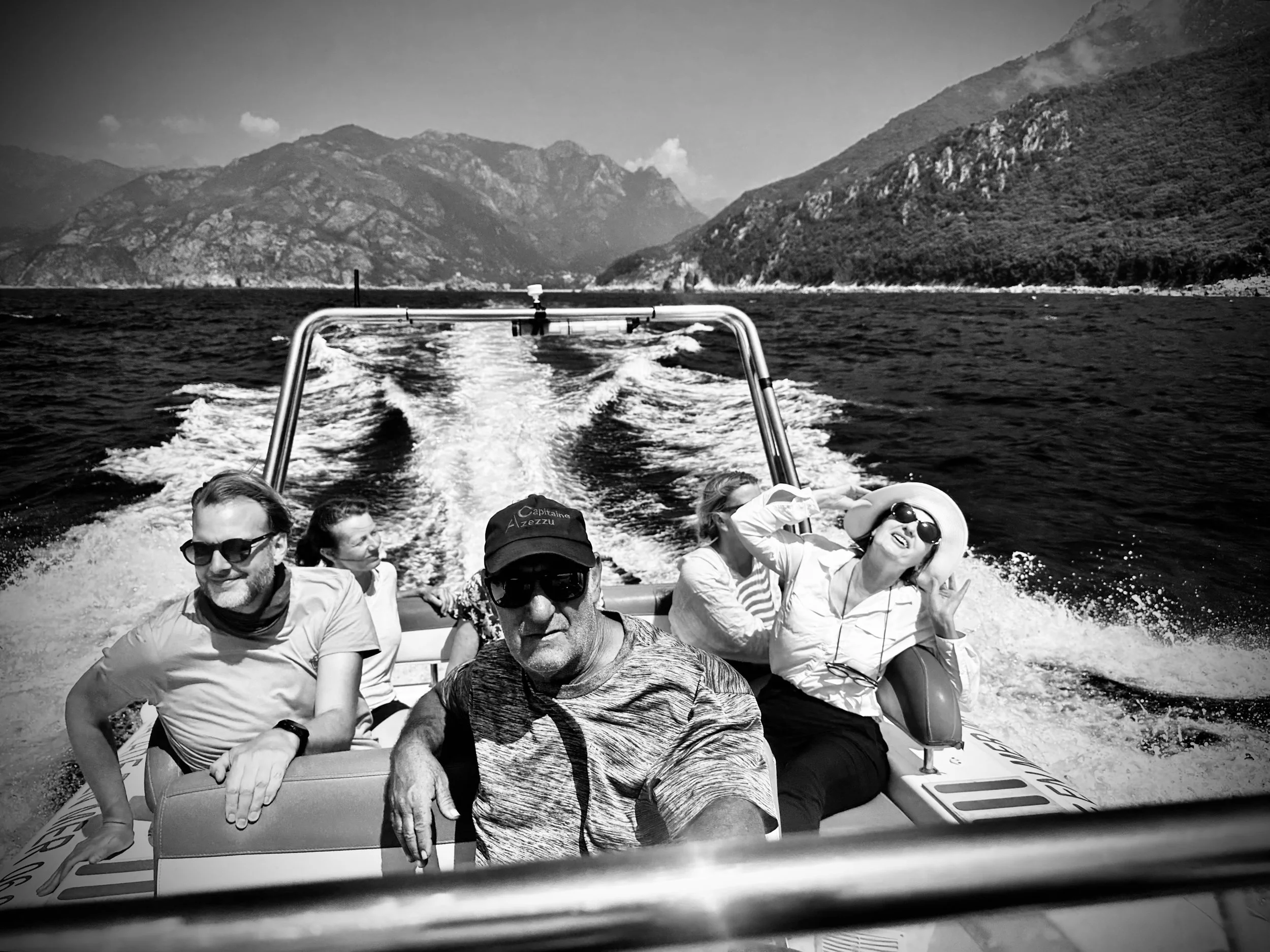 Group of five people enjoying a boat ride on a lake surrounded by mountains, with wake behind the boat and clear sky.
