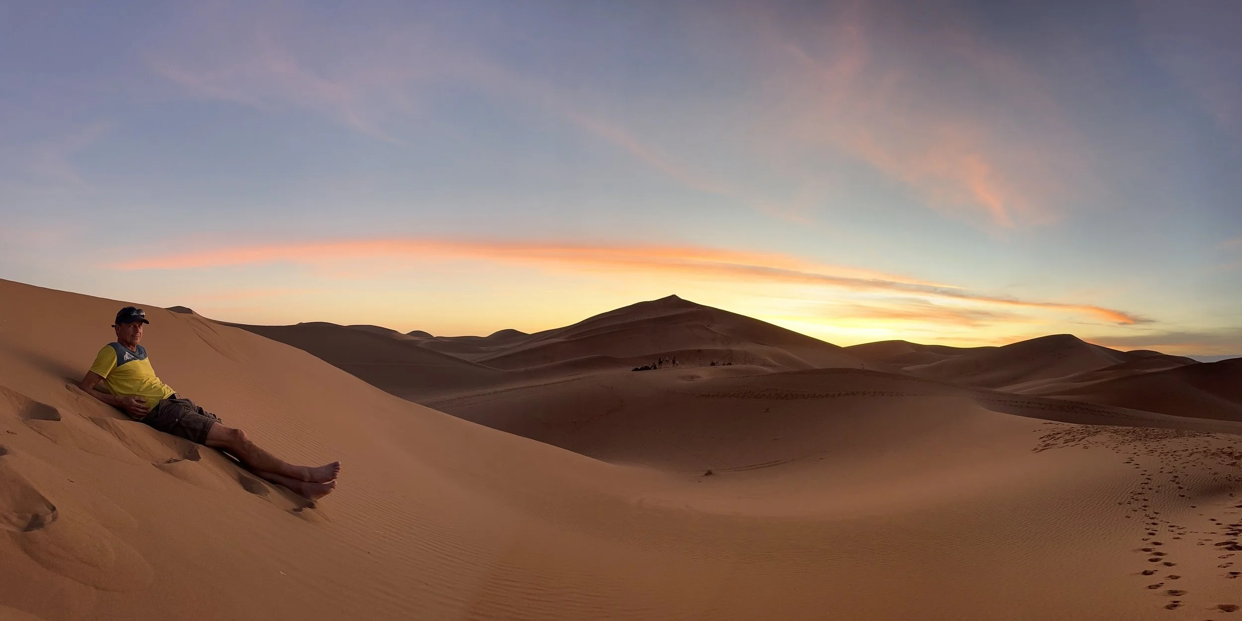 A man sitting on a sand dune in a desert at sunset, with footprints leading towards him and distant dunes and mountains in the background.