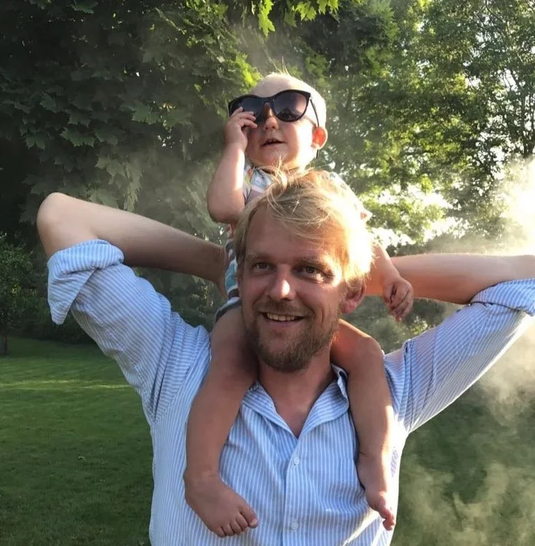 A man with blonde hair and a beard carrying a young child with sunglasses on his shoulders in a park with green trees and grass.