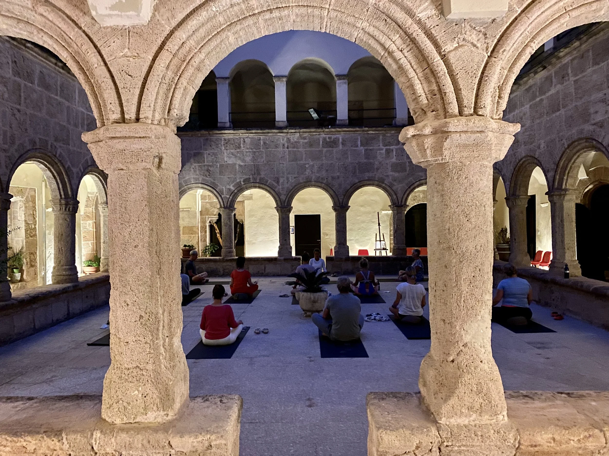 People practicing yoga in a circular stone courtyard with arches at dusk, illuminated by soft lighting.
