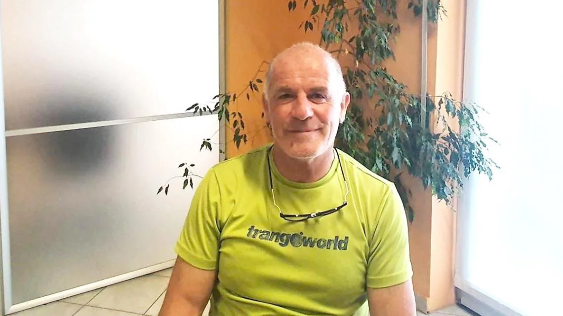 A smiling middle-aged man wearing a lime green t-shirt and glasses hanging around his neck, sitting indoors near a potted plant.