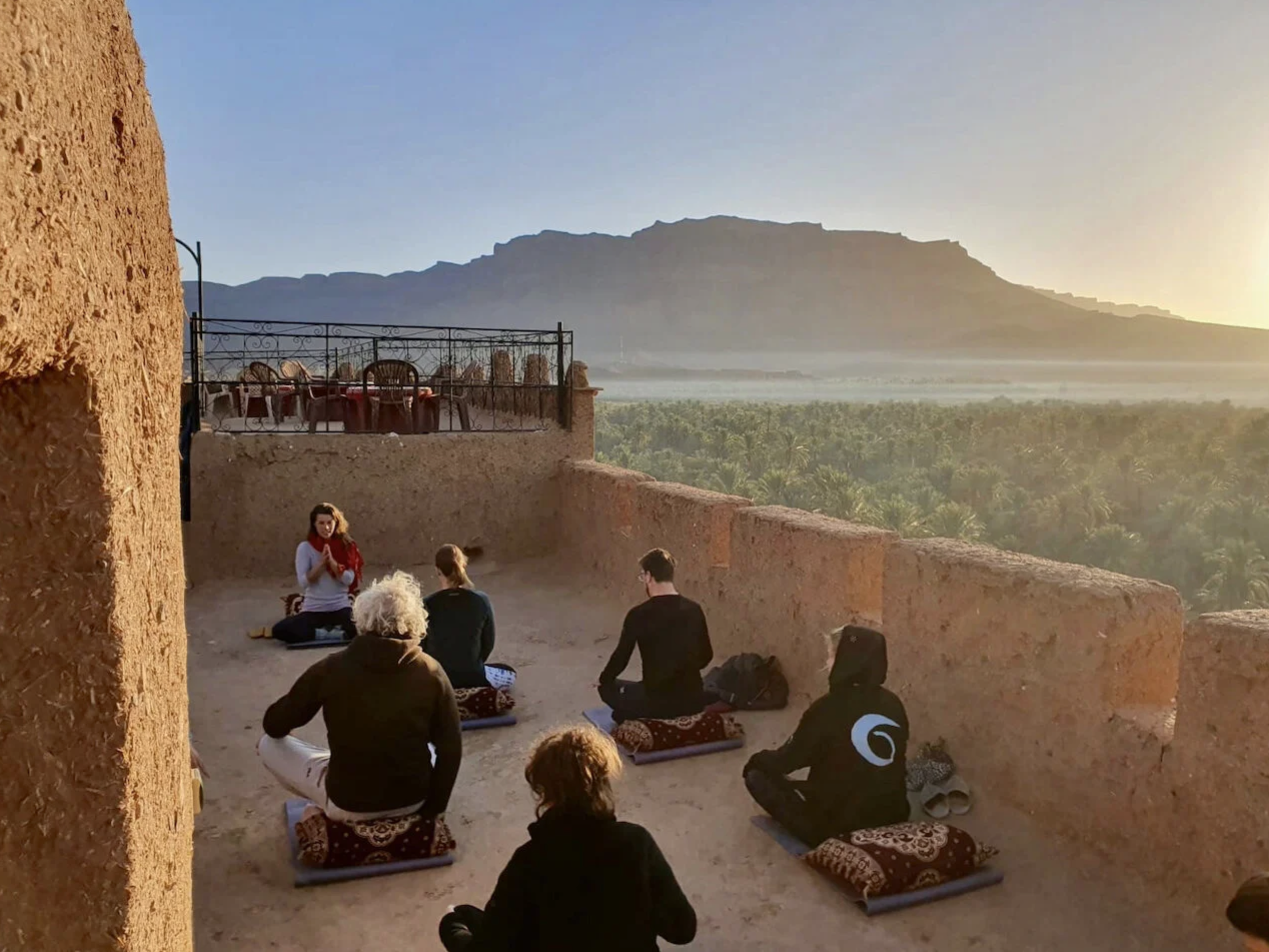 Group of people practicing yoga on a rooftop terrace at sunset, overlooking a lush green landscape and mountains in the background.