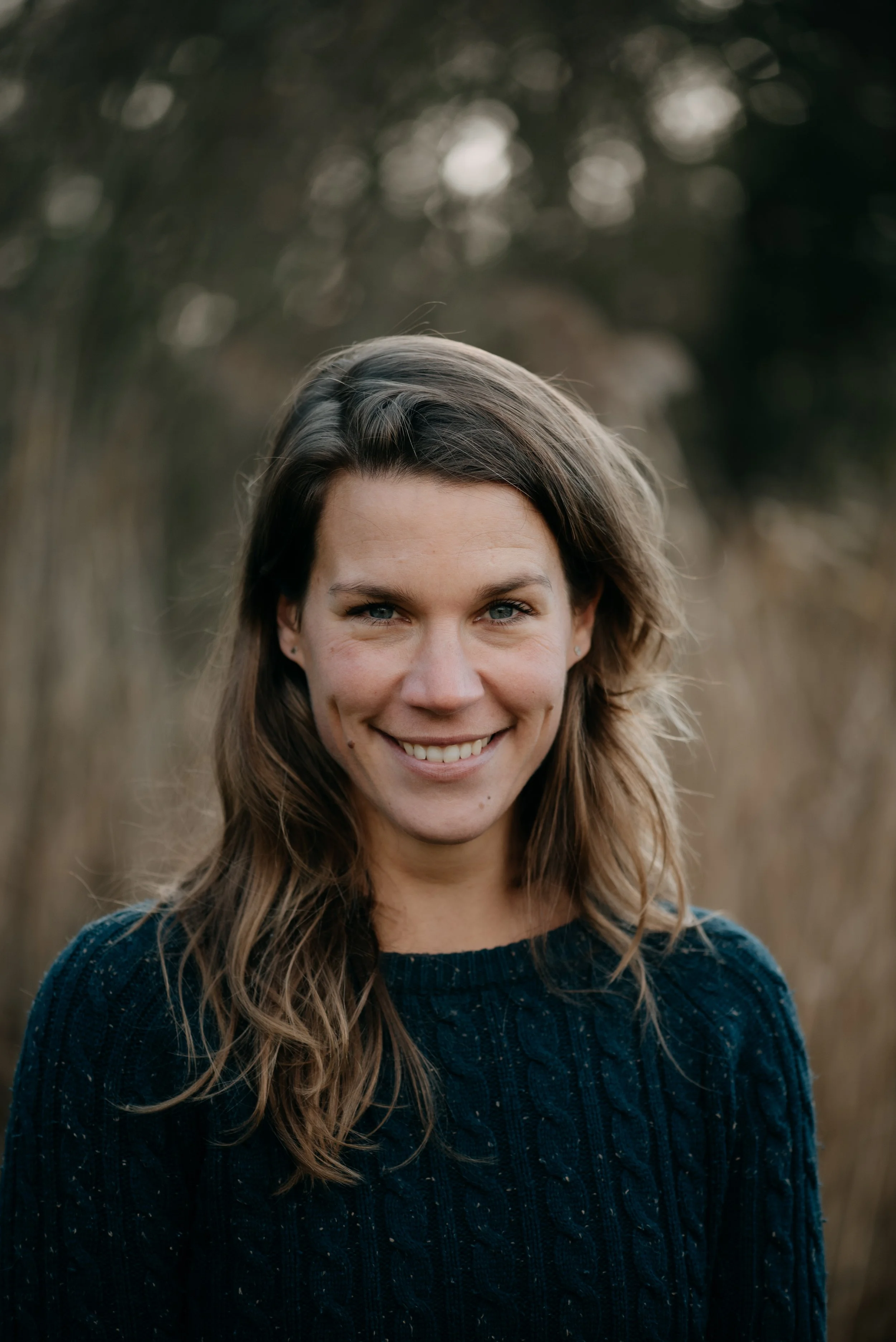 A young woman with long, wavy brown hair and blue eyes, smiling outdoors on a fall day, wearing a dark blue sweater, with blurred trees in the background.