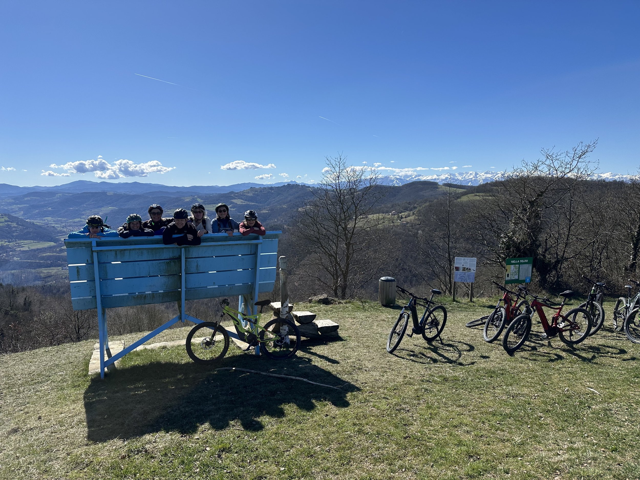 Group of seven people wearing helmets, riding bikes, and standing behind a blue wooden viewing platform in a mountainous landscape with clear blue sky and snow-capped peaks in the background.