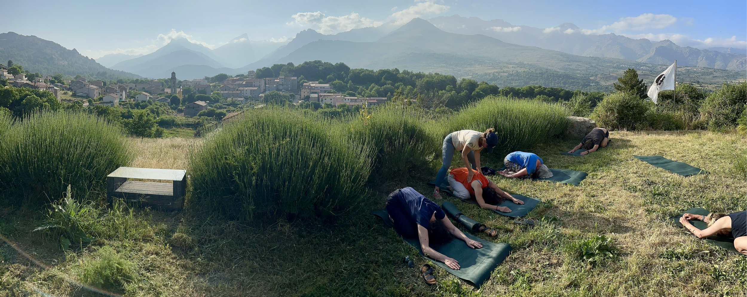 Group of people practicing outdoor yoga on mats in a scenic mountain landscape with greenery, mountains in the background, and a flag in the distance.