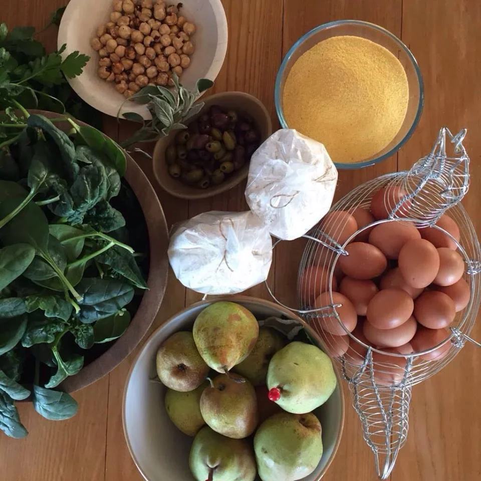 A collection of fresh produce and baking ingredients on a wooden table, including eggs in a wire basket, apples in a bowl, chickpeas, green olives, herbs, and a glass bowl of yellow cornmeal.