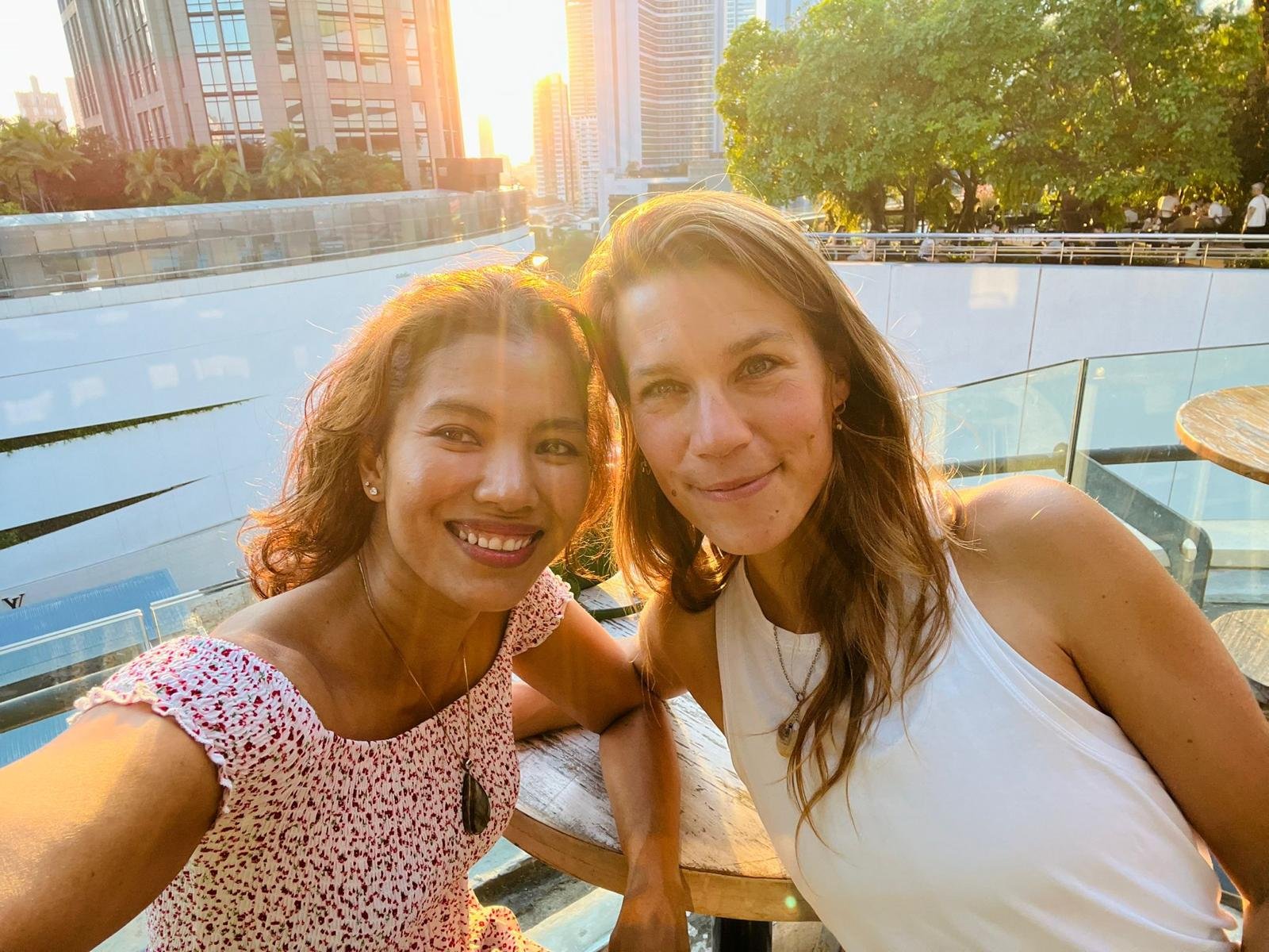 Two women taking a selfie on a rooftop patio during sunset in a city with tall buildings and green trees.