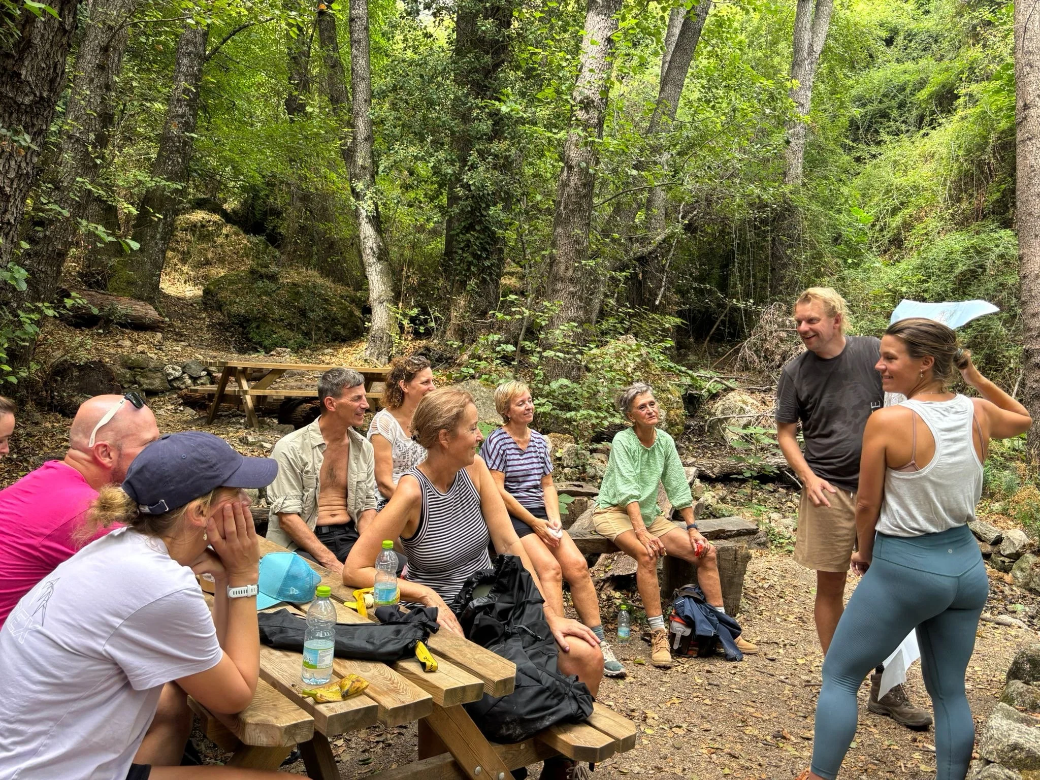 Group of people sitting on a wooden picnic table and rocks in a forest, listening and watching two people standing and talking.