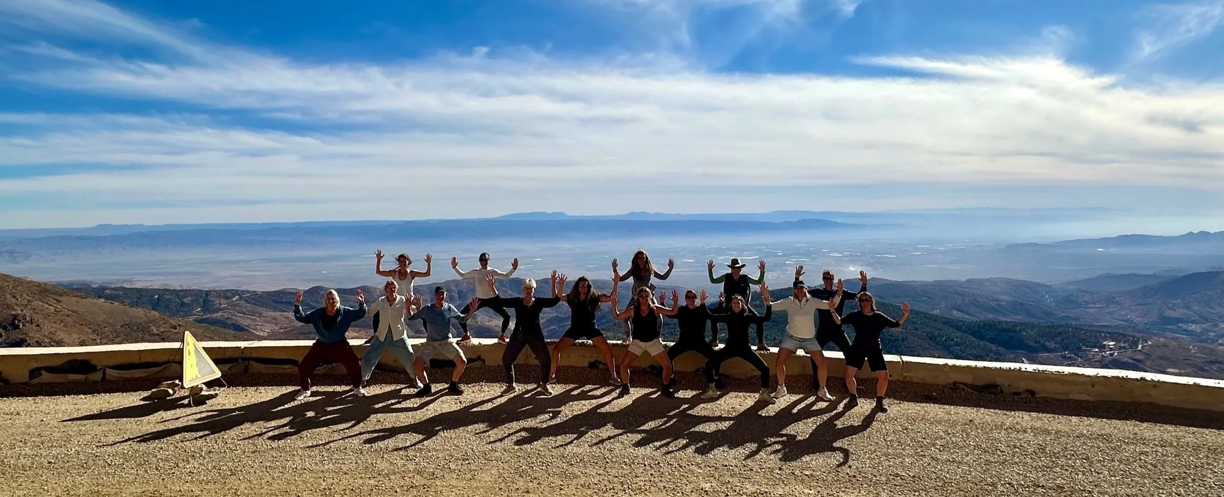 Group of fifteen people standing on a mountain overlook with a scenic view of valleys and mountains in the background. The group is posing with arms raised, casting long shadows on the ground.