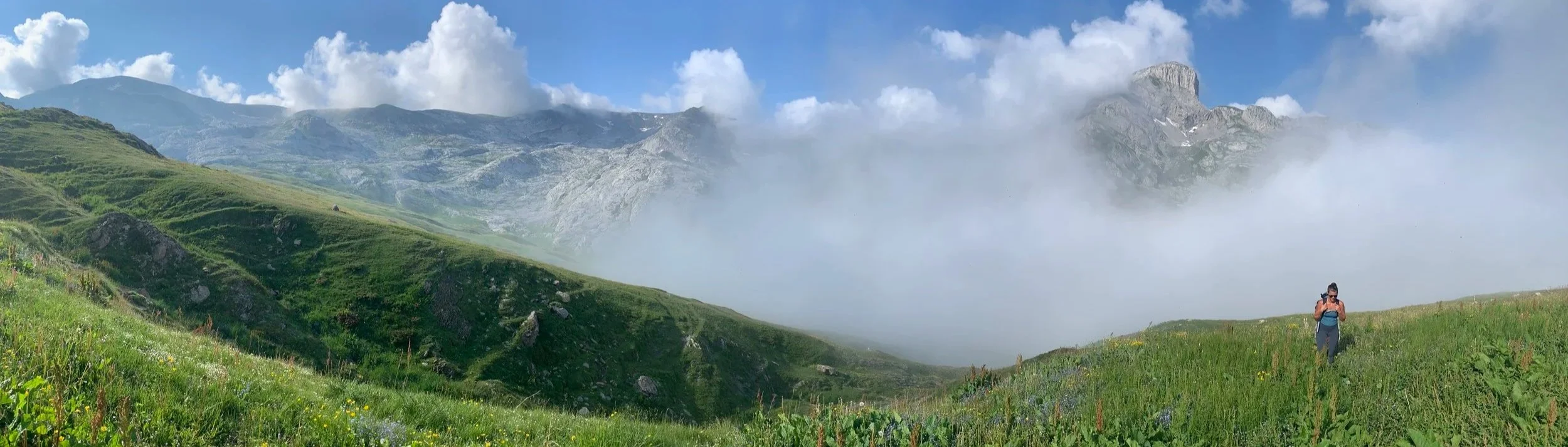 A person hiking on a grassy hill in a mountainous area with clouds and fog in the background.