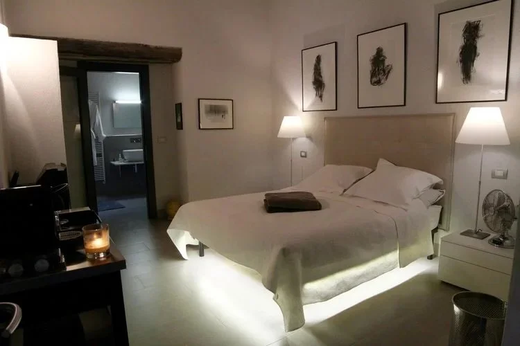 A cozy hotel bedroom with a bed, nightstands, lamps, and wall art, illuminated by soft lighting.