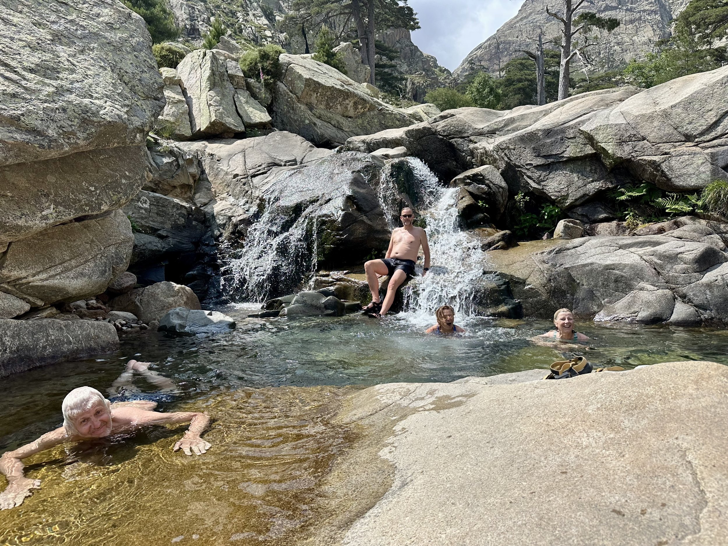 Four people enjoying a natural rock pool with a small waterfall in a rocky outdoor setting, surrounded by large rocks, trees, and mountains in the background.