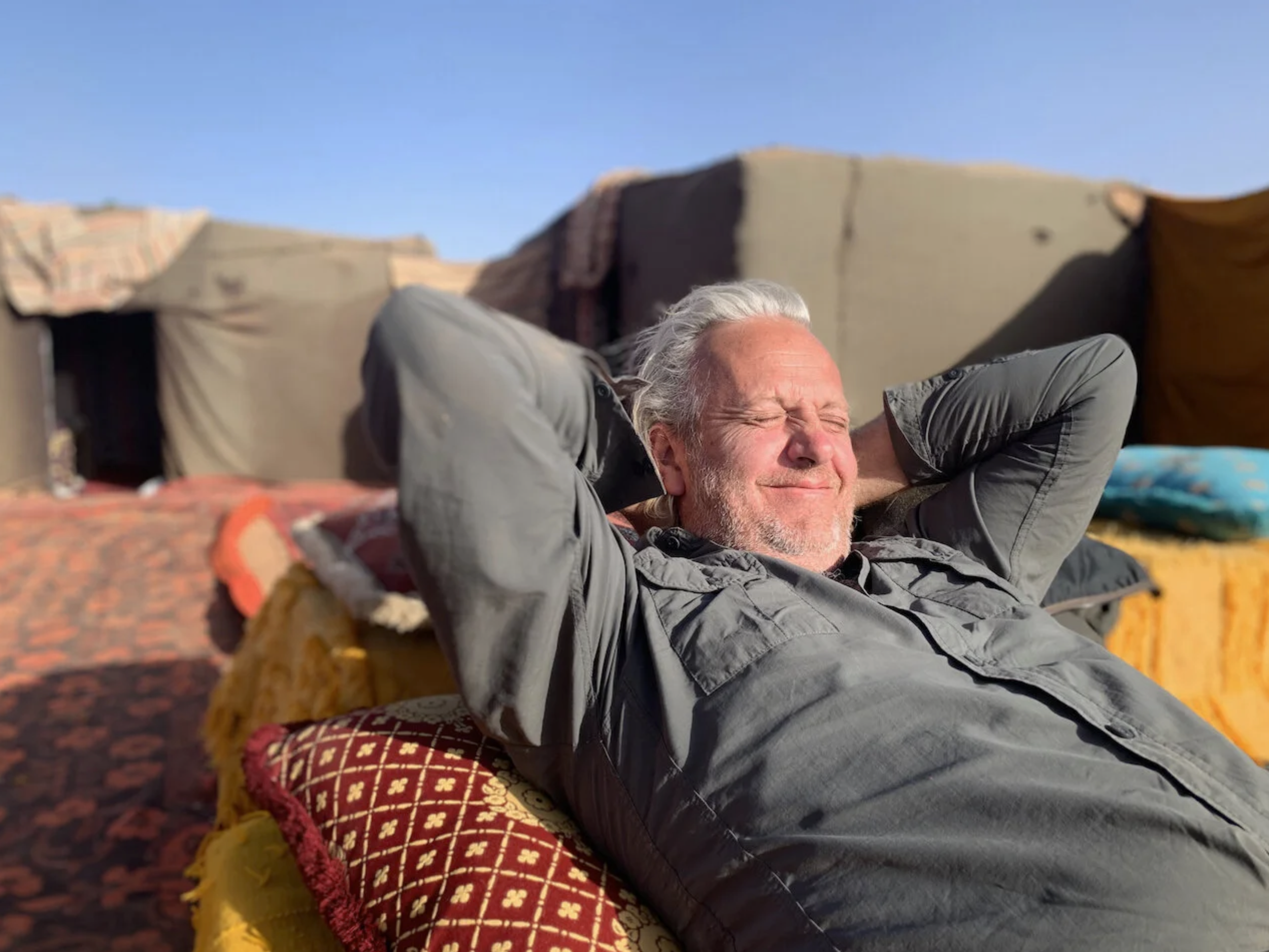 An elderly man relaxing outdoors on a sunny day, lying back with arms behind his head and eyes closed. He is seated on a decorative cushion on the ground, with colorful textiles and blankets around him. The background features makeshift tents or structures under a clear blue sky.