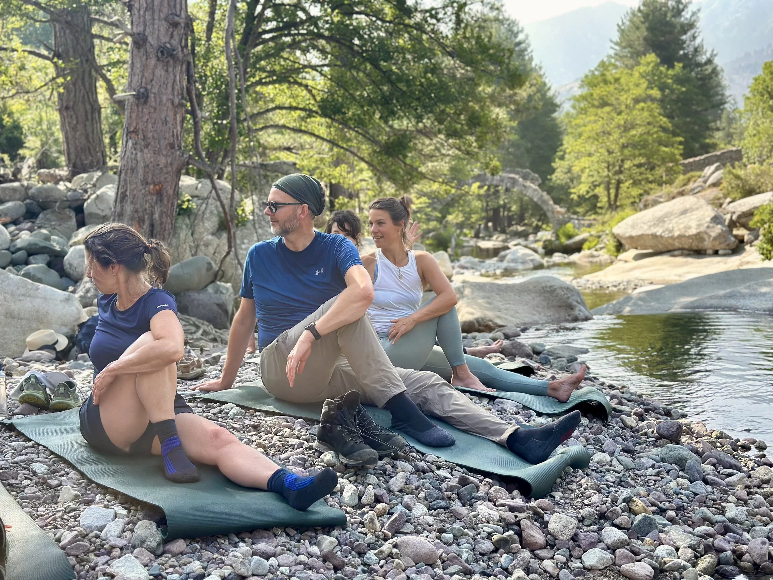 Group of people doing yoga and stretching on yoga mats on a rocky riverbank in a forest with water and trees.