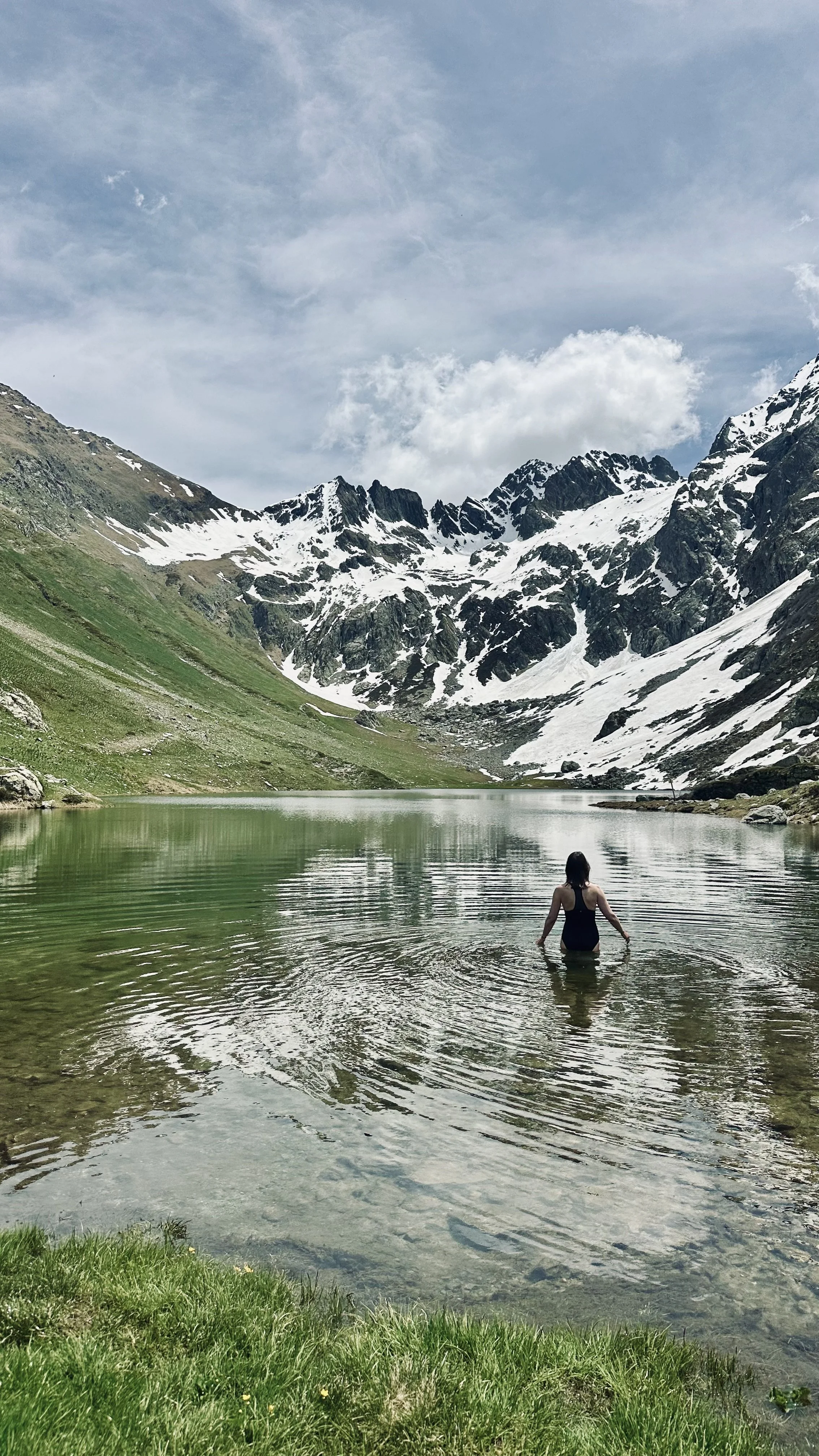 A woman wading in a lake surrounded by mountains with snow and green grass, under partly cloudy sky.