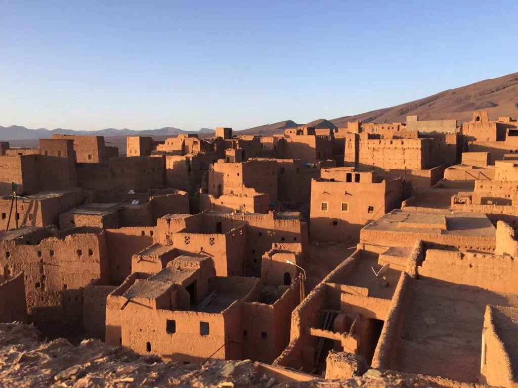 Adobe-style mud-brick buildings in a desert village at sunset with mountains in the background.