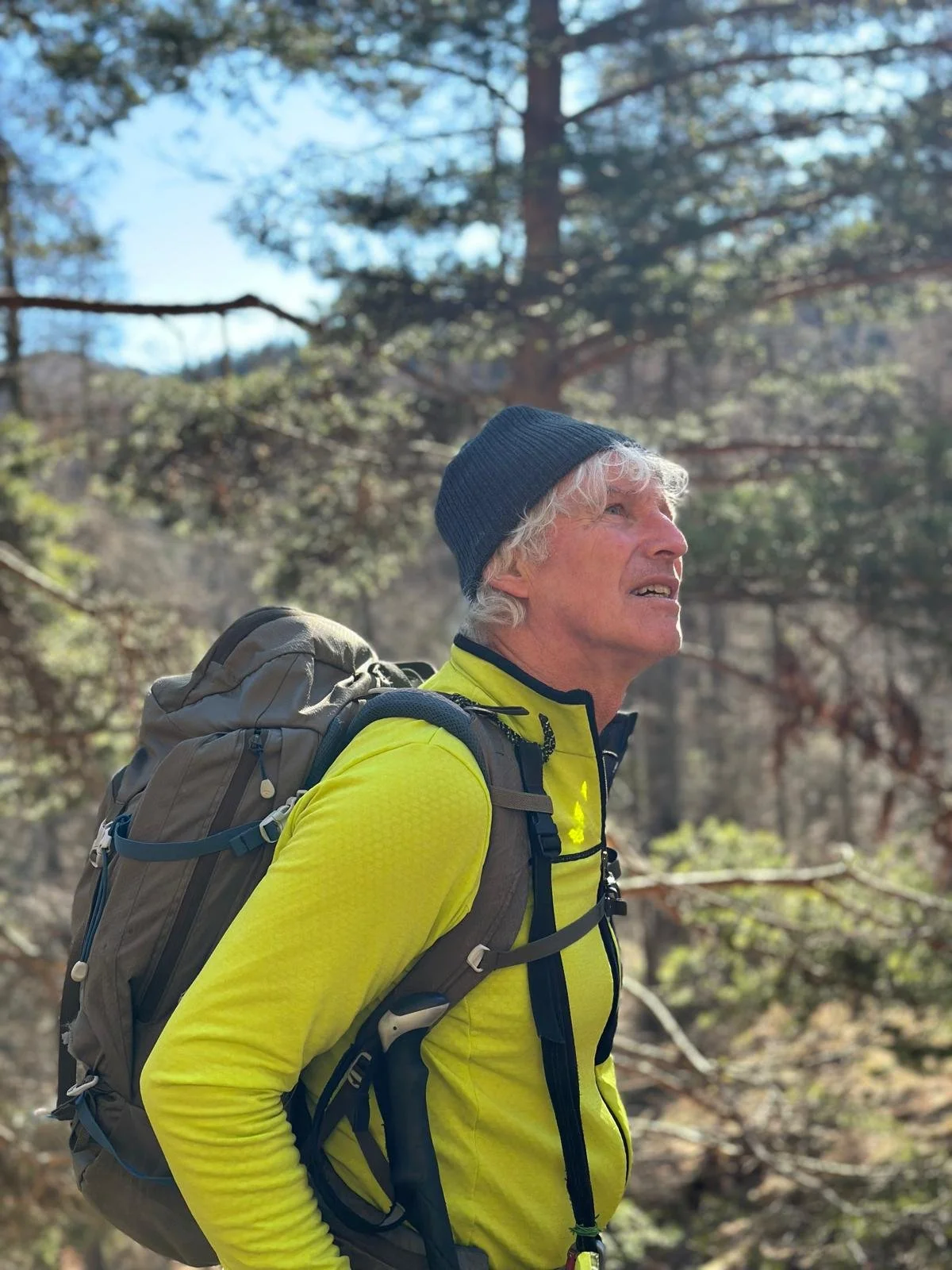 Older man with gray hair, wearing a blue beanie and a bright yellow jacket, hiking outdoors with a large backpack in a forested area.
