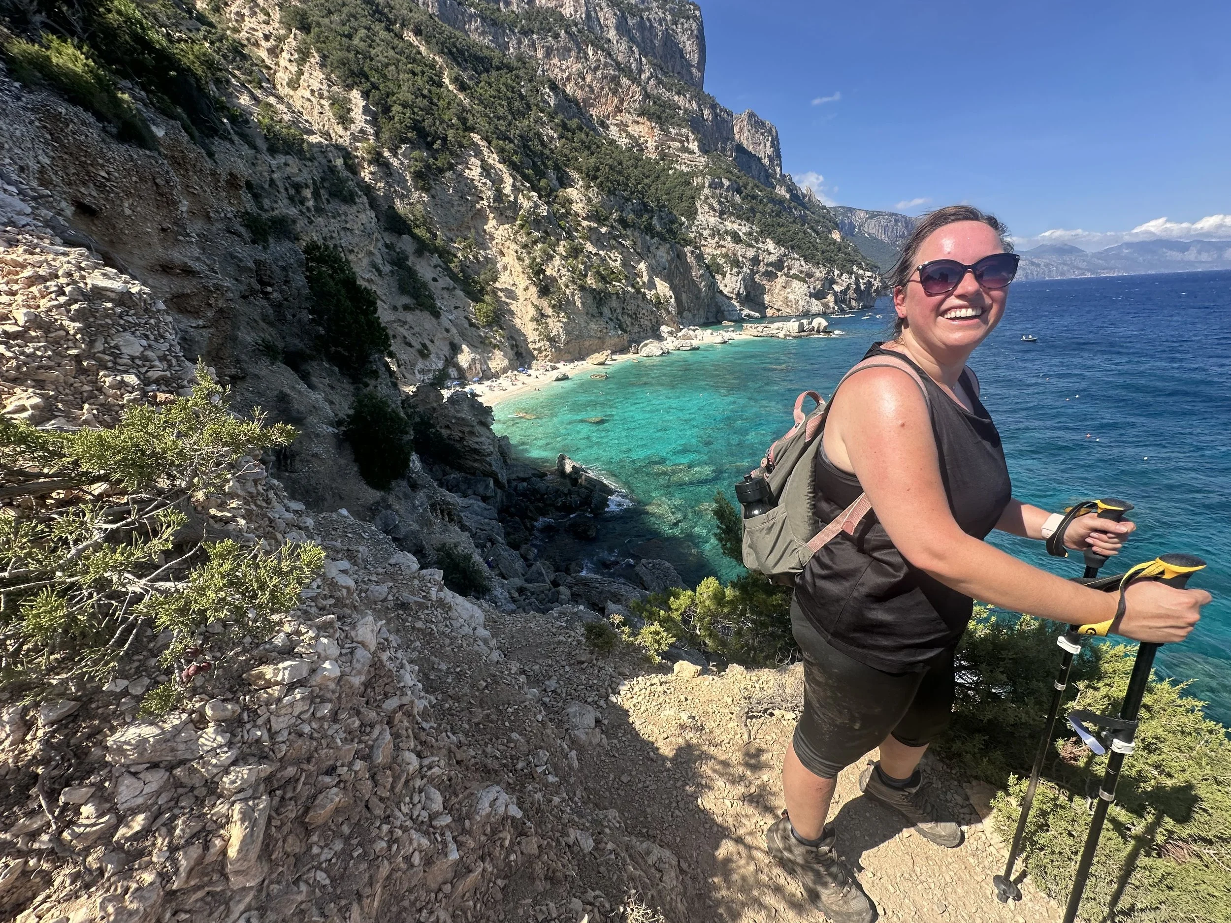 A woman smiling with sunglasses, wearing a black sleeveless top and black pants, hiking along a cliffside trail with a backpack and trekking poles, overlooking a turquoise bay with rocky cliffs and trees under a clear blue sky.