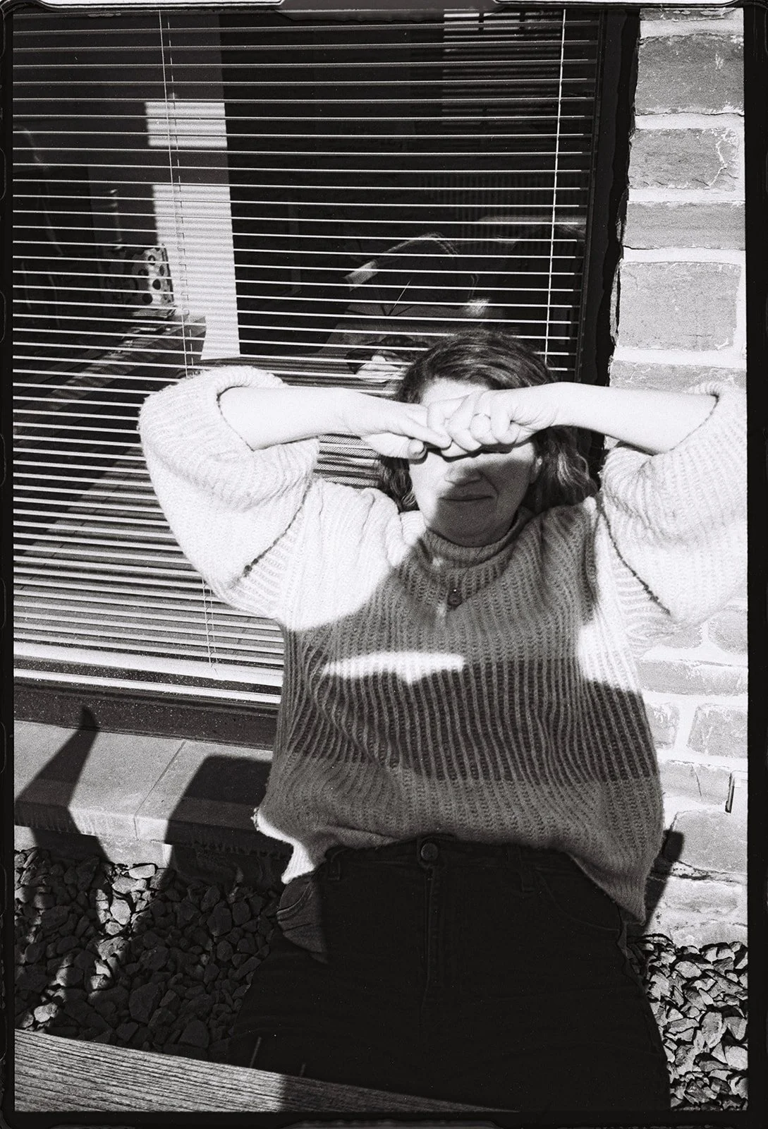 Een persoon zit buiten op een bank, droeg een trui en heeft haar handen voor haar gezicht, de persoon kijkt naar beneden. Ze schuift haar handen onder haar neus, alsof ze zich verstopt of zich beschaamd voelt.