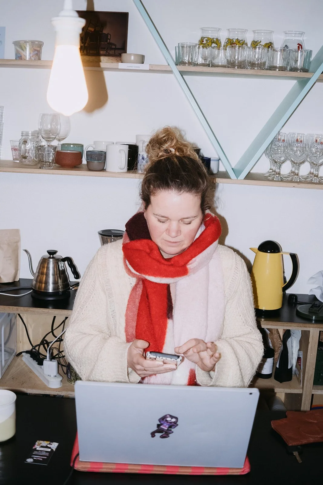 Vrouw met een rood en beige sjaal die op haar telefoon kijkt, zit voor een laptop in een keuken of eetruimte.