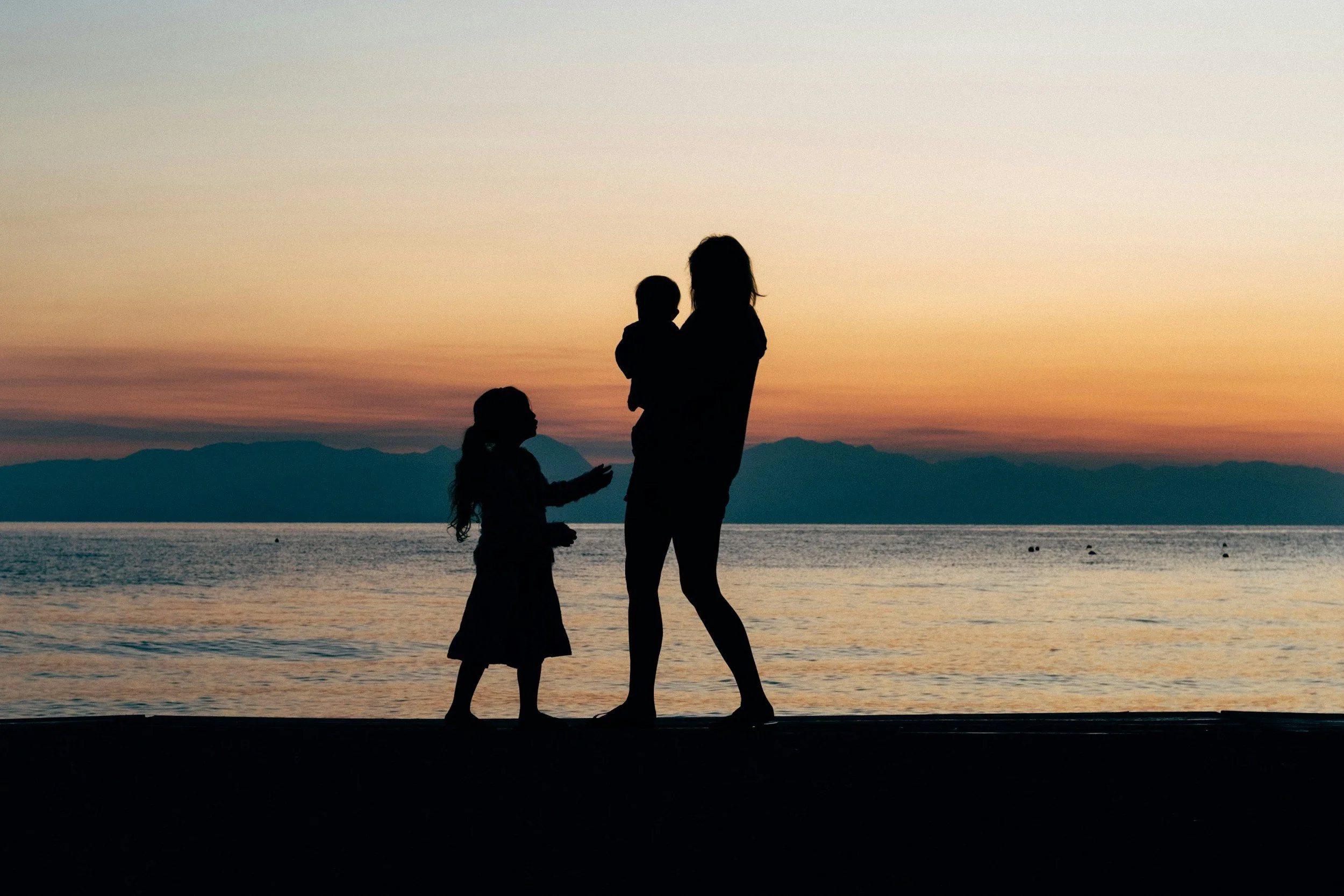 Silhouettes of a woman, a young girl, and a small child by the water during sunset with mountains in the background.