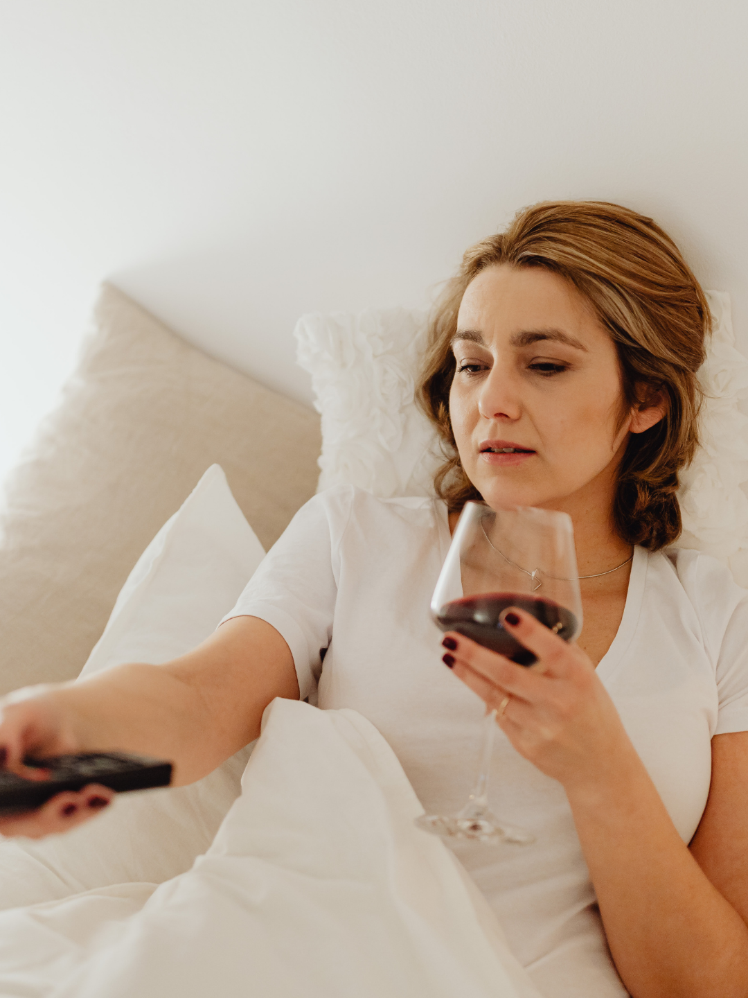 Bild zeigt Frau, die es sich im Bett mit Wein und Fernbedienung gemütlich gemacht hat und ihr Business einfach mal nebenbei laufen lässt
