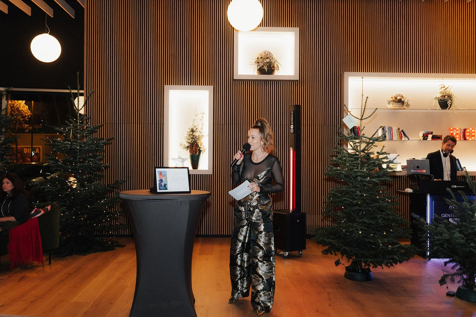 A woman in elegant black and gold patterned outfit is speaking into a microphone at an indoor event with Christmas trees and modern decor.