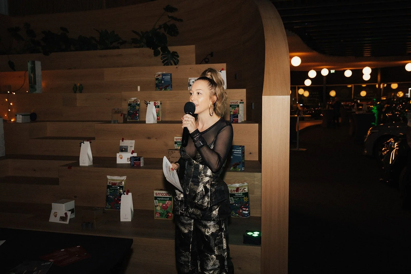 Frau mit blondem Pferdeschwanz, Mikrofon und Moderationskarten in den Händen, die in einem gediegenen Ambiente spricht.