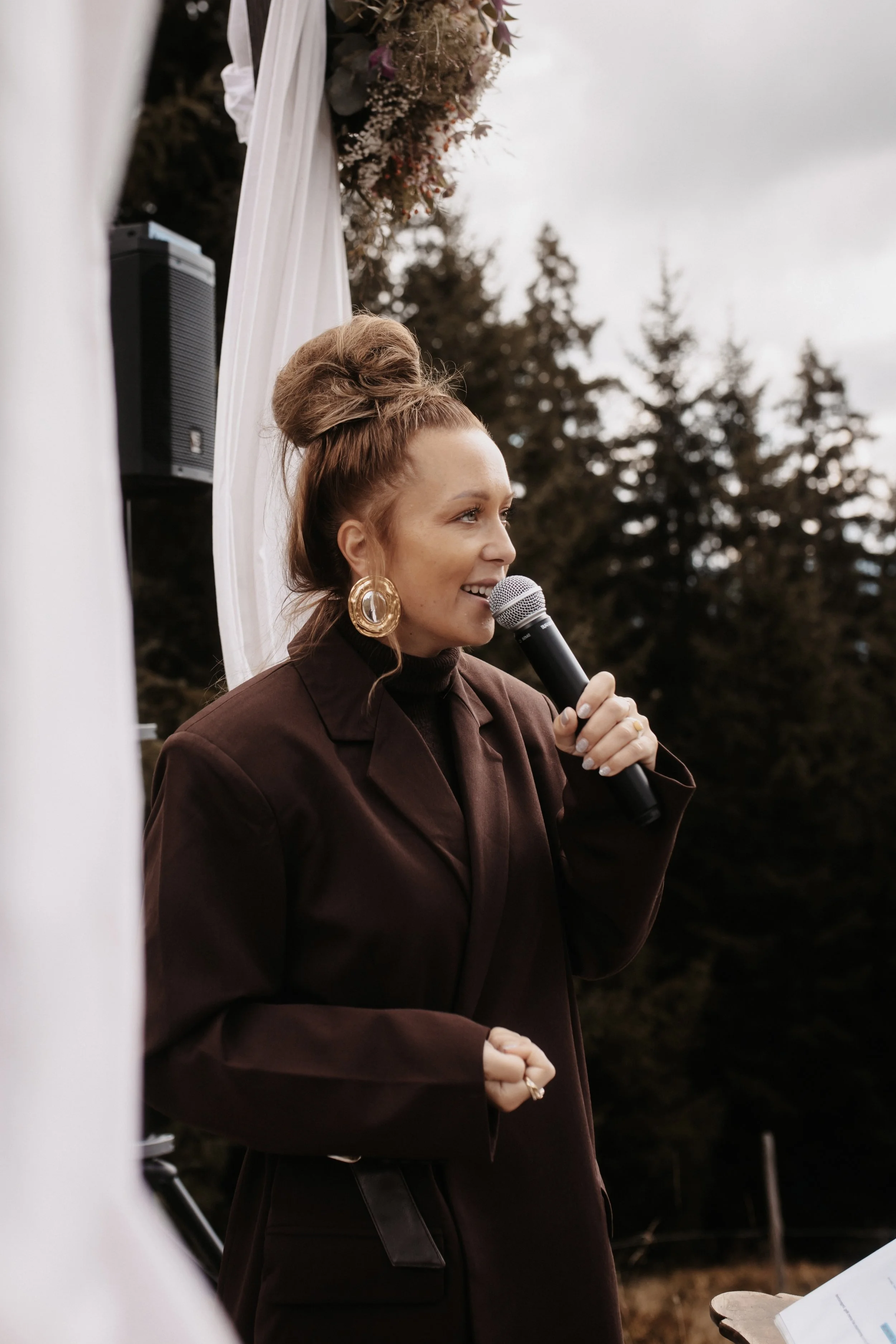 A woman with large gold earrings and a voluminous updo hairstyle speaking into a microphone outdoors during a cloudy day, with trees in the background and white drapery decorated with flowers behind her.