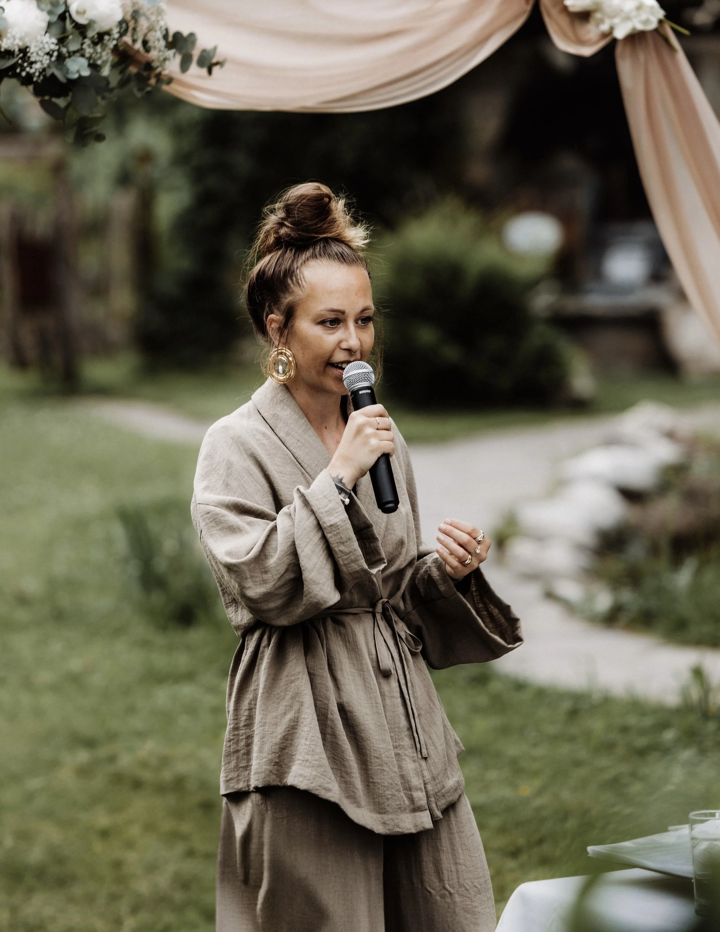 Frau mit messy Dutt und Mikrofon in der Hand, die für ein Hochzeitspaar spricht.