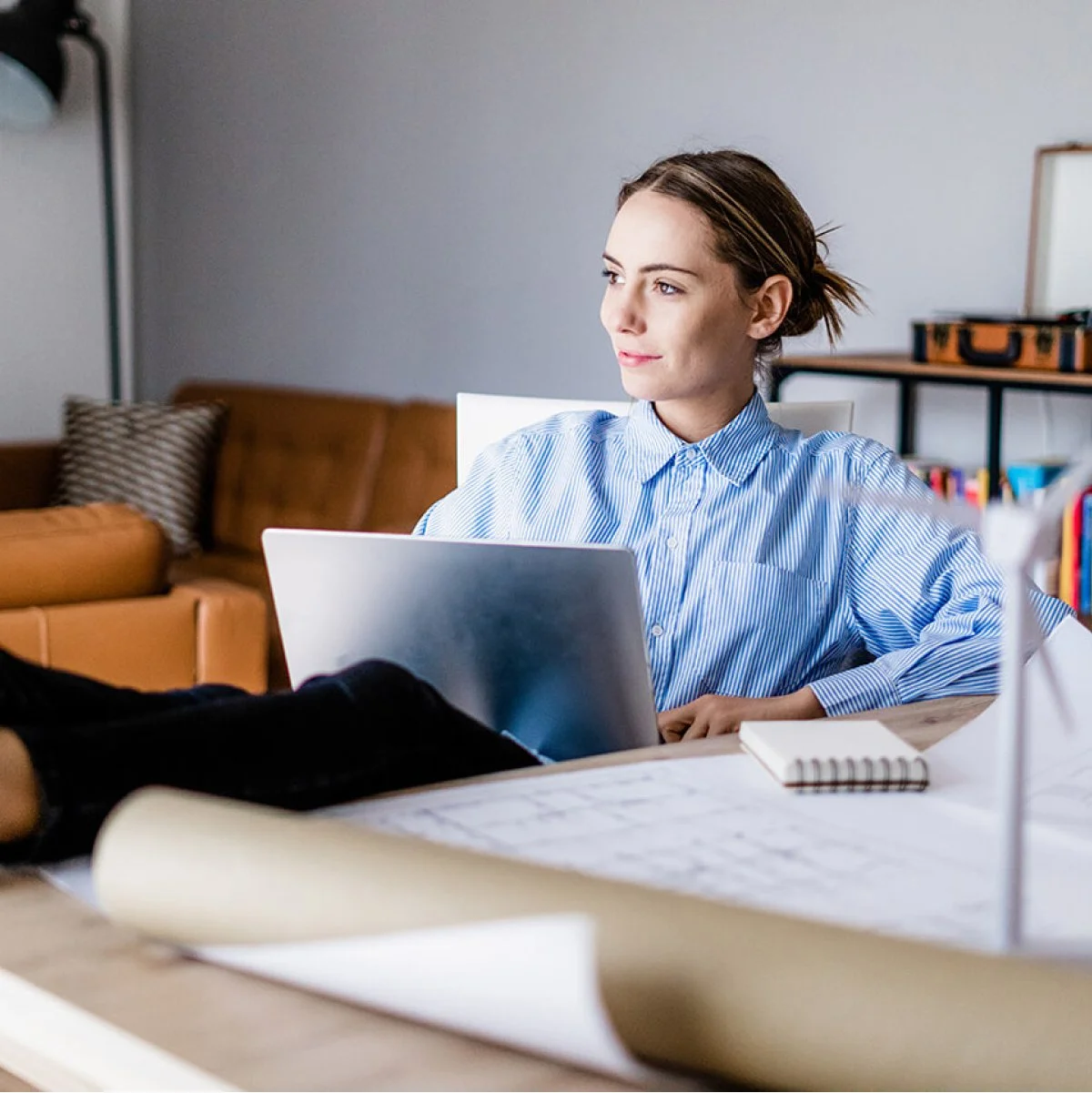 Junge Frau arbeitet am Laptop in einem Wohnzimmer, das mit Büchern, einem Notizbuch und einem Sofa ausgestattet ist.