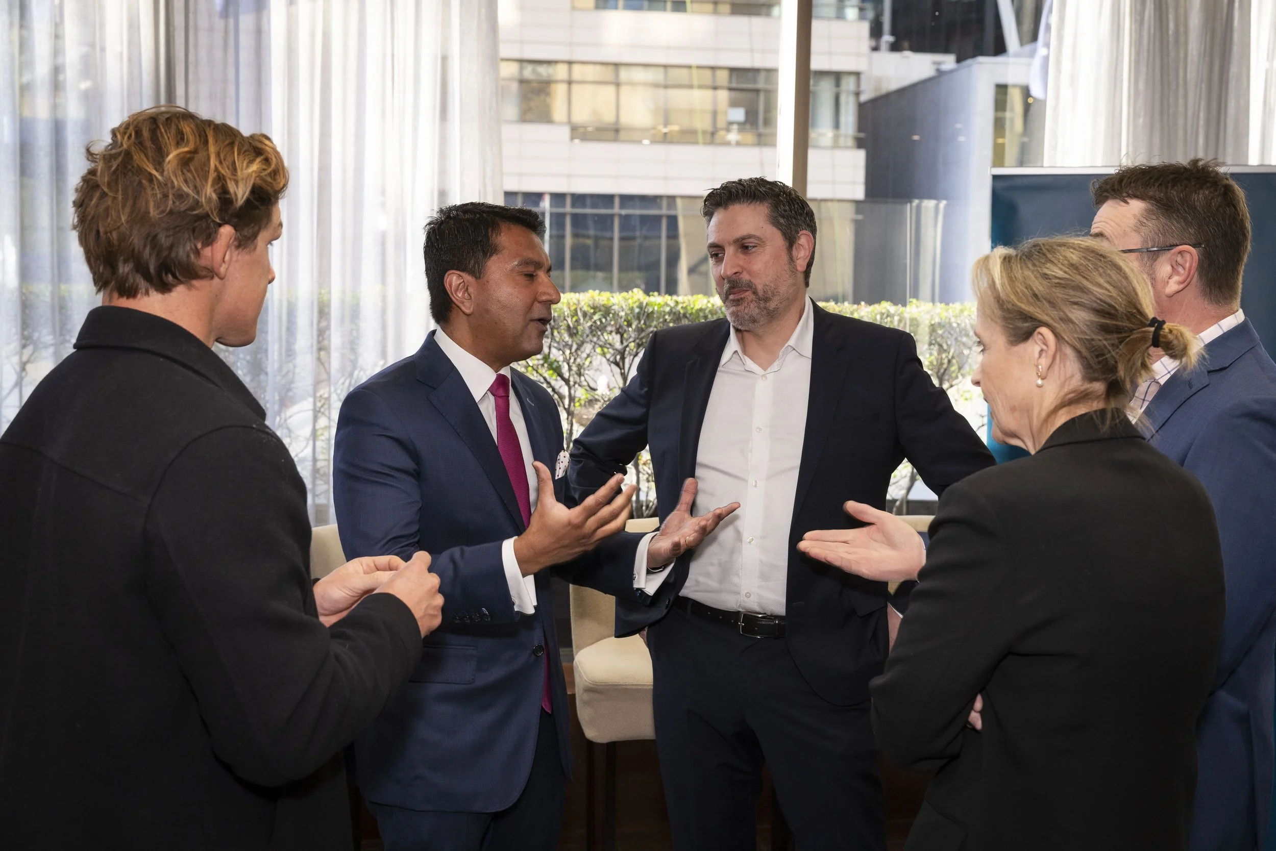 Group of five business professionals engaged in discussion, standing in a modern office with large windows.