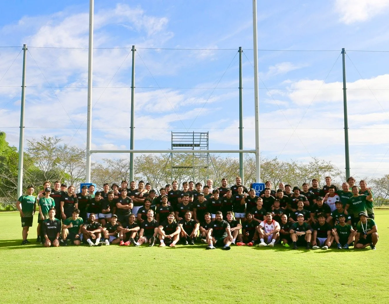 A large rugby team posing for a group photo on a rugby field with goal posts and trees in the background, some team members wearing green jerseys and others in black and white jerseys, under a partly cloudy sky.