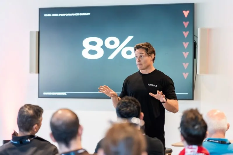 A presenter stands in front of a screen displaying '8%' during a high-performance summit, speaking to an audience of seated attendees.