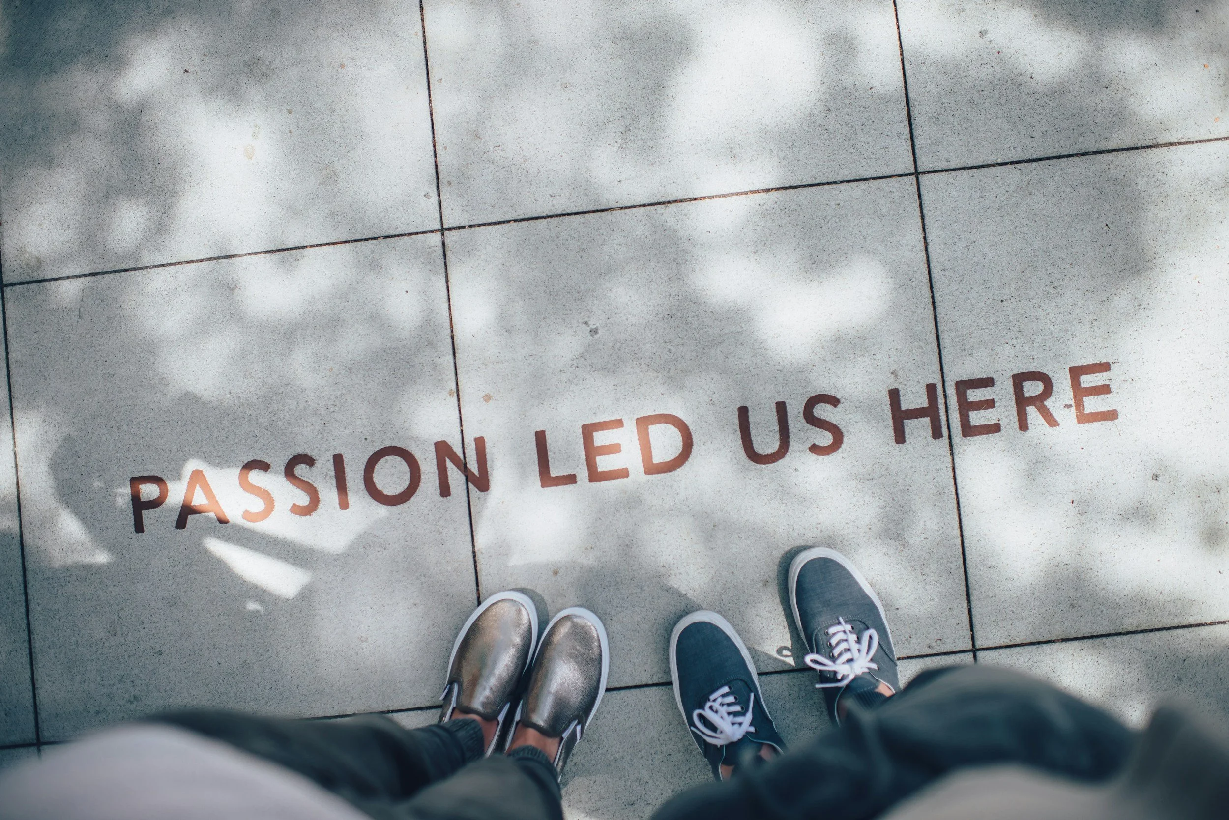 View of two people standing on gray tiled pavement with shadow of tree, looking down at the text 'PASSION LED US HERE' painted on the ground
