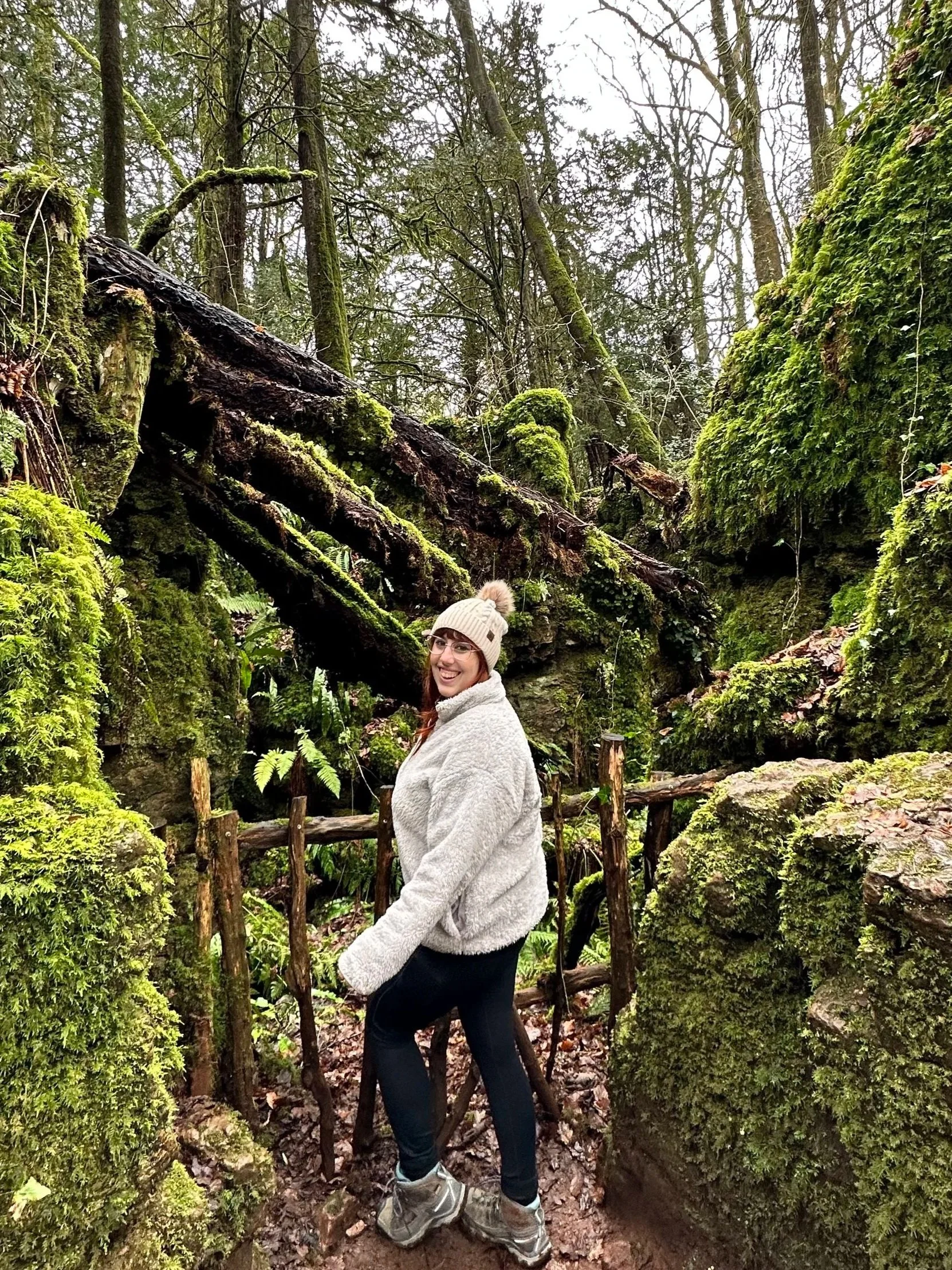 A woman smiling in a beige knit hat and light gray fleece jacket standing on a forest trail surrounded by moss-covered rocks and fallen logs.