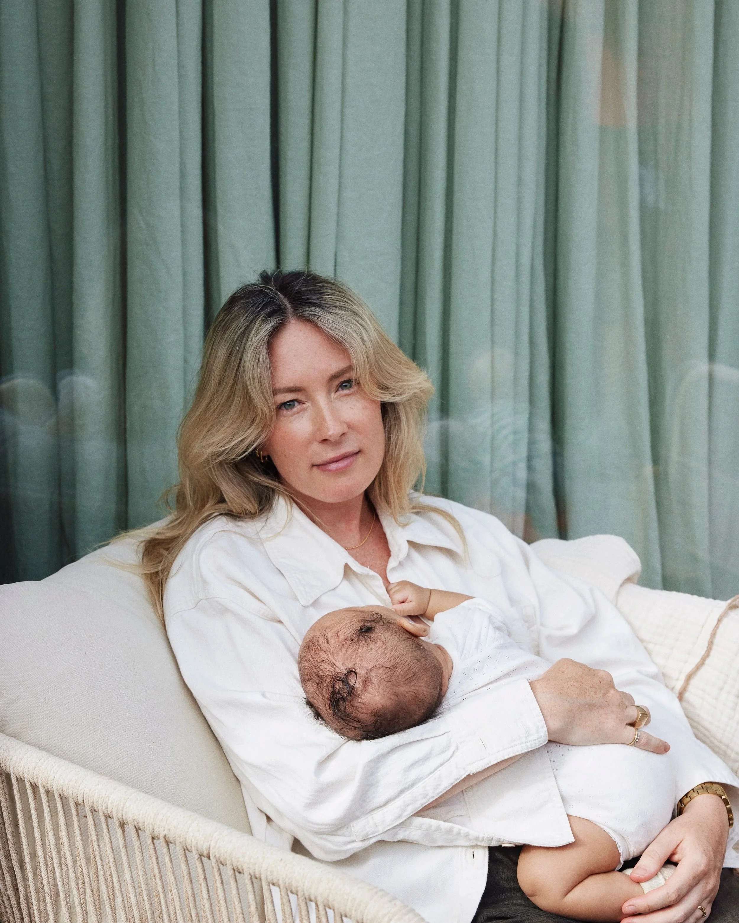Woman in a white shirt breastfeeding a baby on a couch in front of green curtains