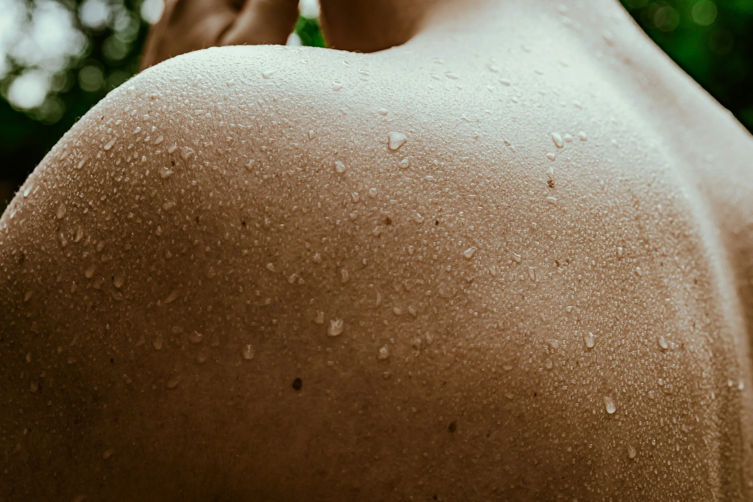 Close-up of a person's shoulder with water droplets on the skin, outdoors with greenery in the background.