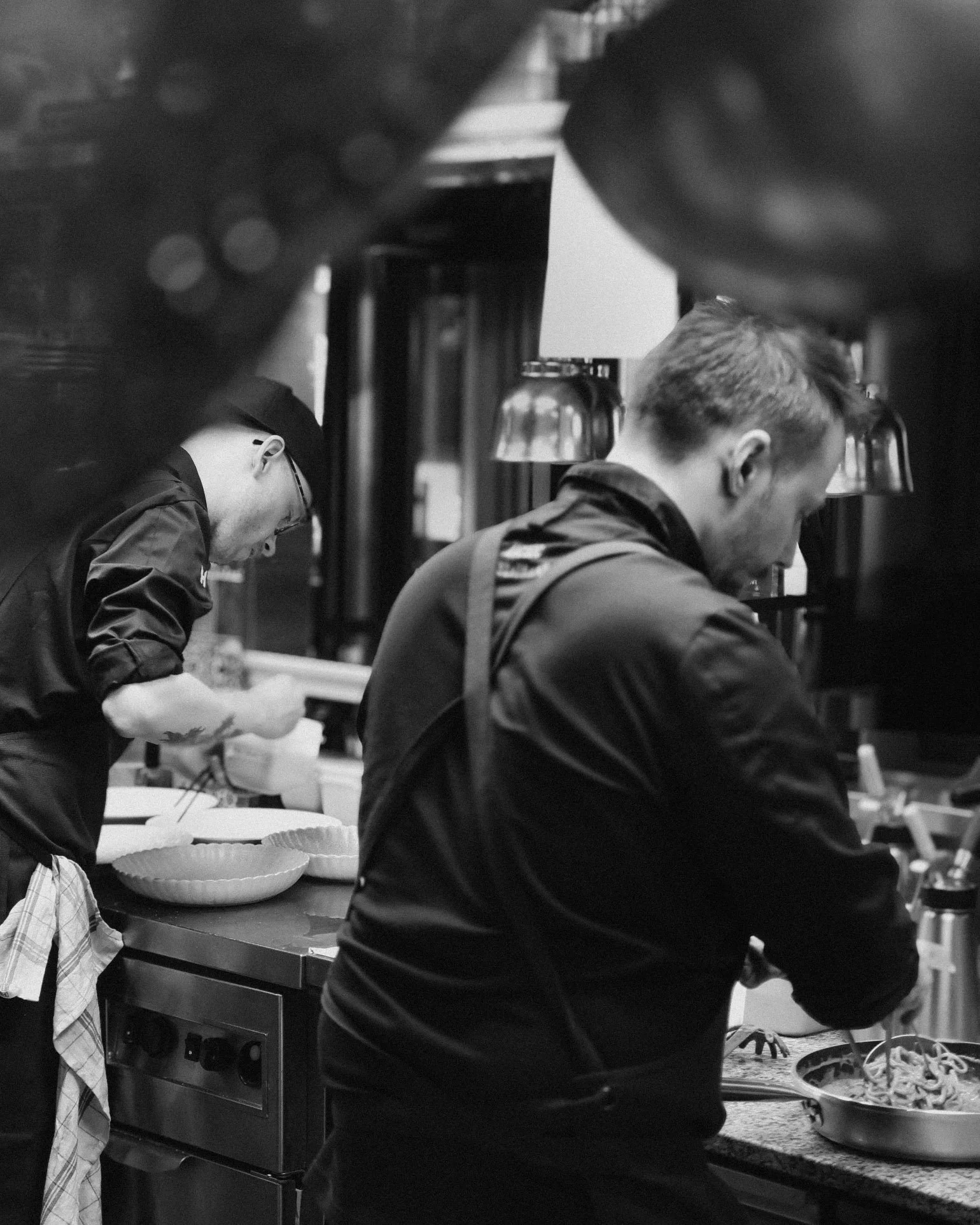 Two chefs in a professional kitchen preparing dishes, seen through a gap in the kitchen's upper cabinets, black and white photo.