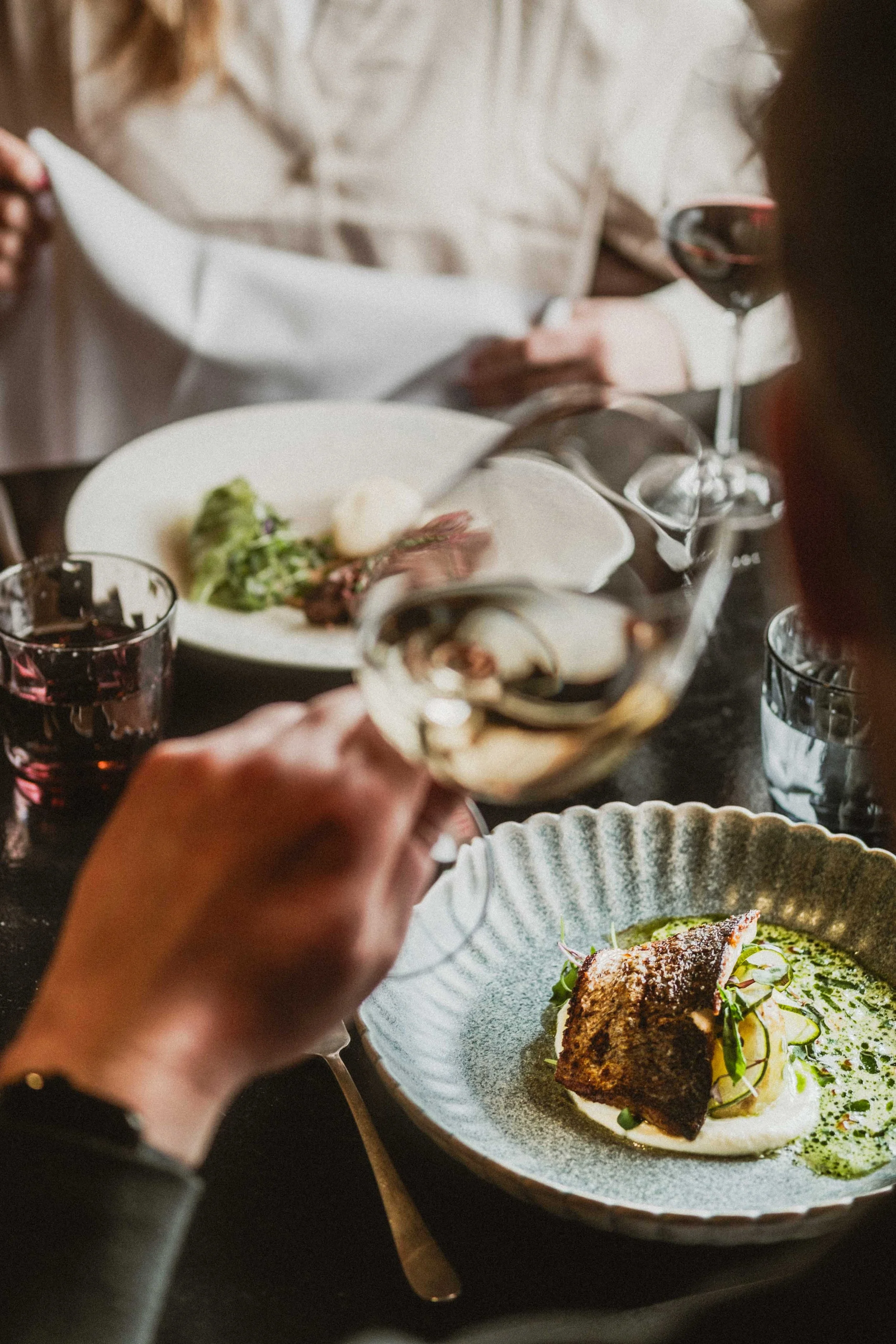 People dining at a restaurant, with one person pouring white wine into a glass over a plate of food that appears to be a gourmet dish garnished with sauce and herbs.