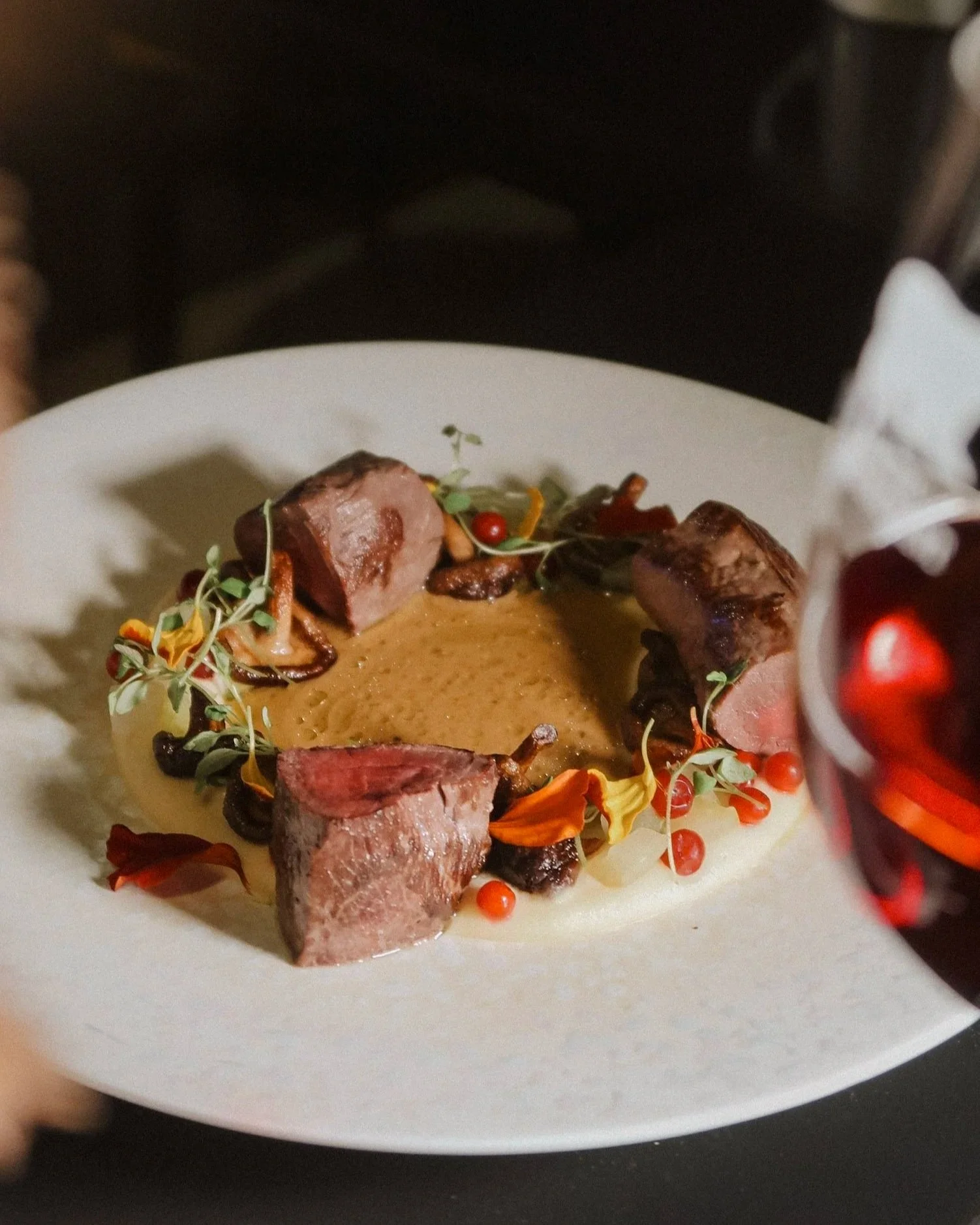 A gourmet dish with neatly sliced seared steak pieces arranged around a creamy sauce or puree, garnished with herbs, microgreens, edible flowers, and small berries, served on a white plate.