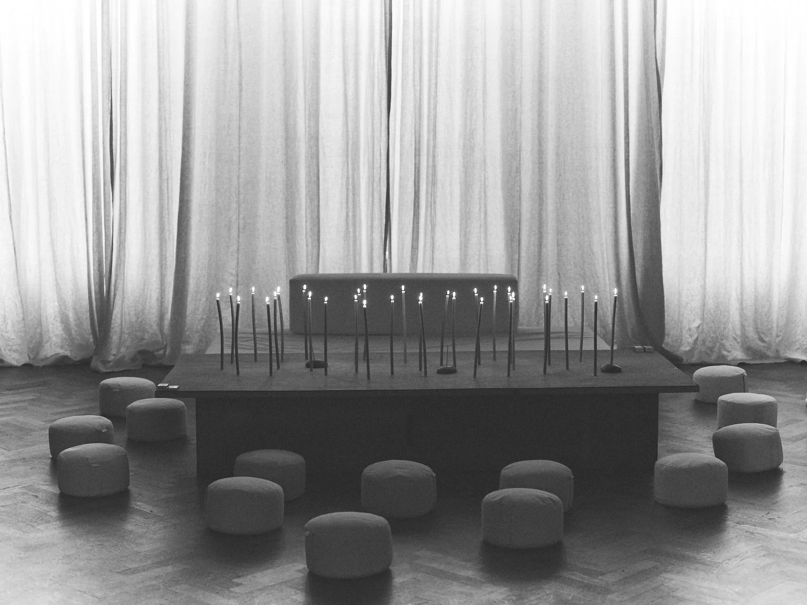 Room with closed curtains, a table in front of the curtains with lit candles, small cushioned seats all around the table on a wooden floor.