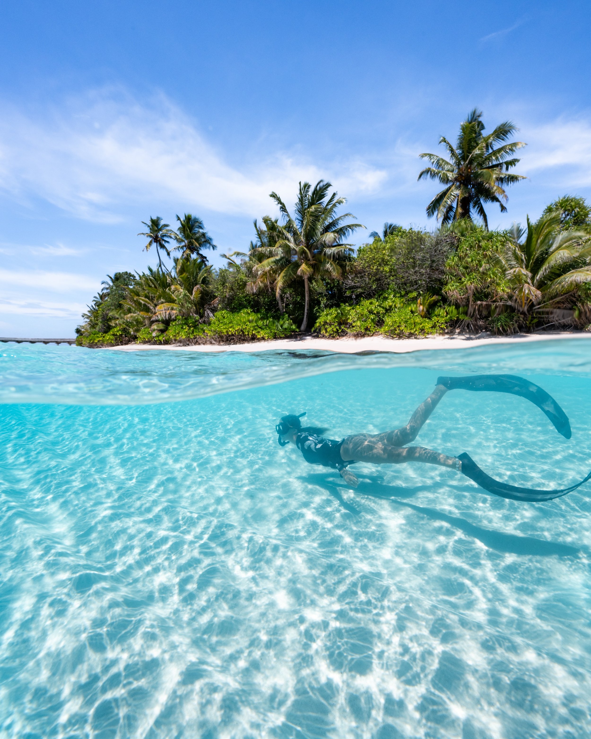 A person snorkeling underwater in clear blue water near a tropical island with palm trees and a bright sky.
