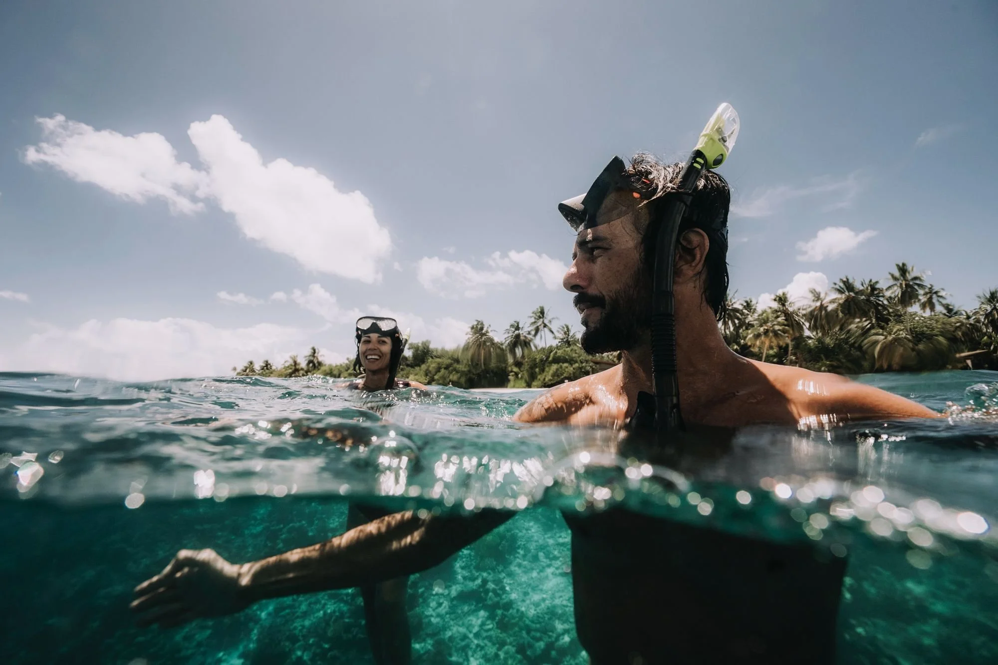 A man and woman snorkeling in clear ocean water near a tropical island with palm trees.