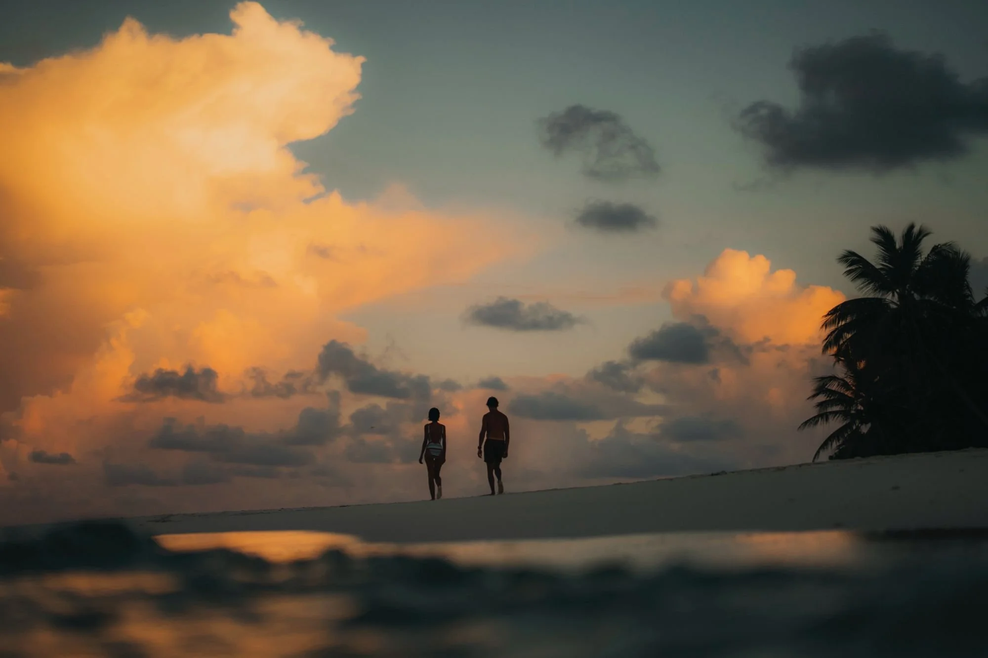 Silhouettes of a man and woman walking on the beach at sunset, with palm trees and clouds in the sky.
