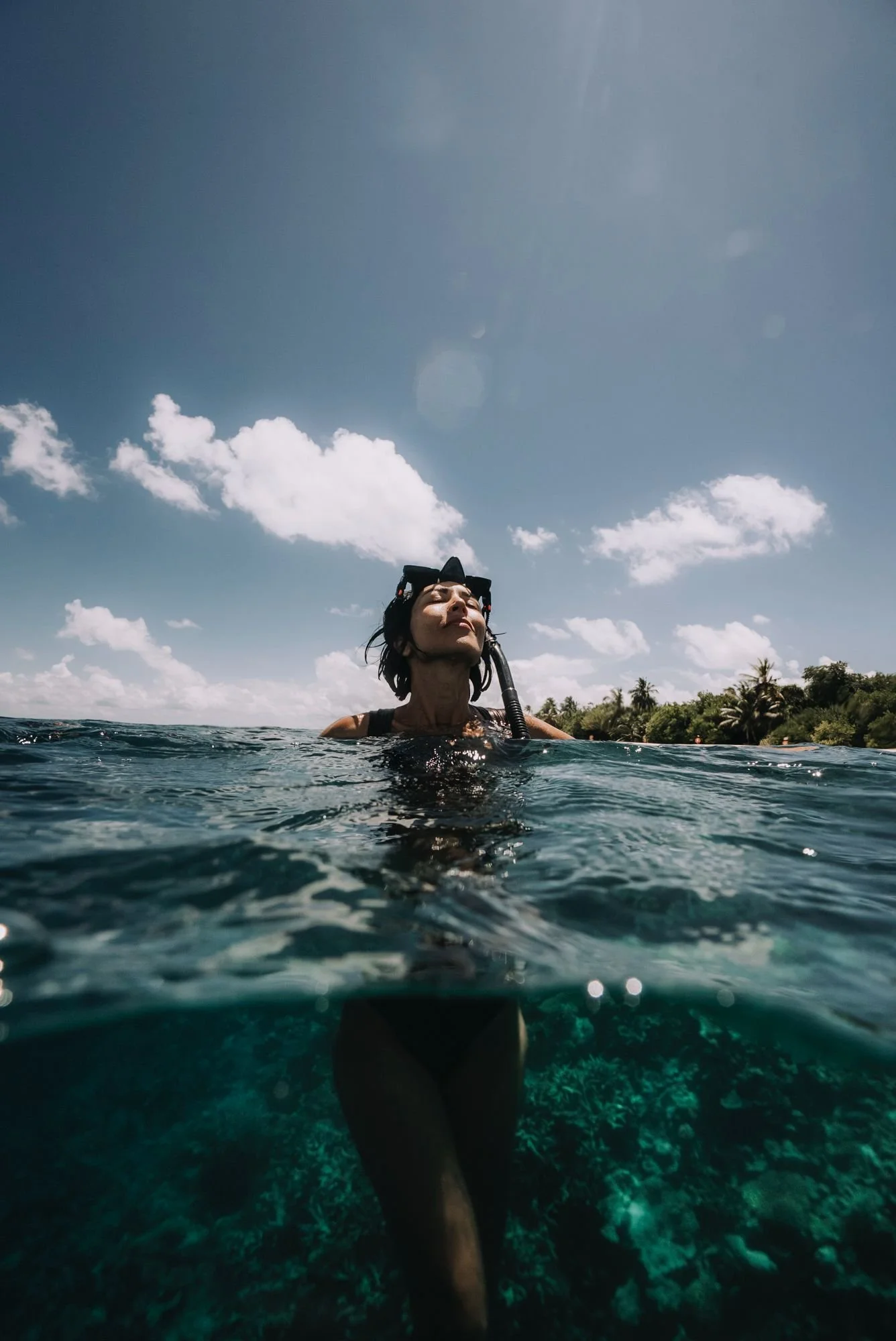 Woman snorkeling in the ocean with her eyes closed, wearing a snorkel and mask, under a partly cloudy sky, with a tropical shoreline in the background.