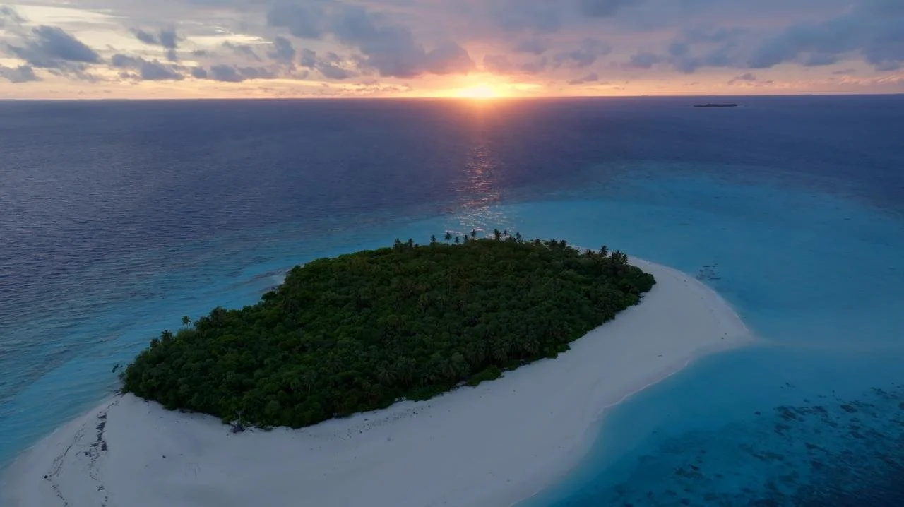 A small, lush green island with a sandy beach in the turquoise ocean, sunset on the horizon with partly cloudy sky