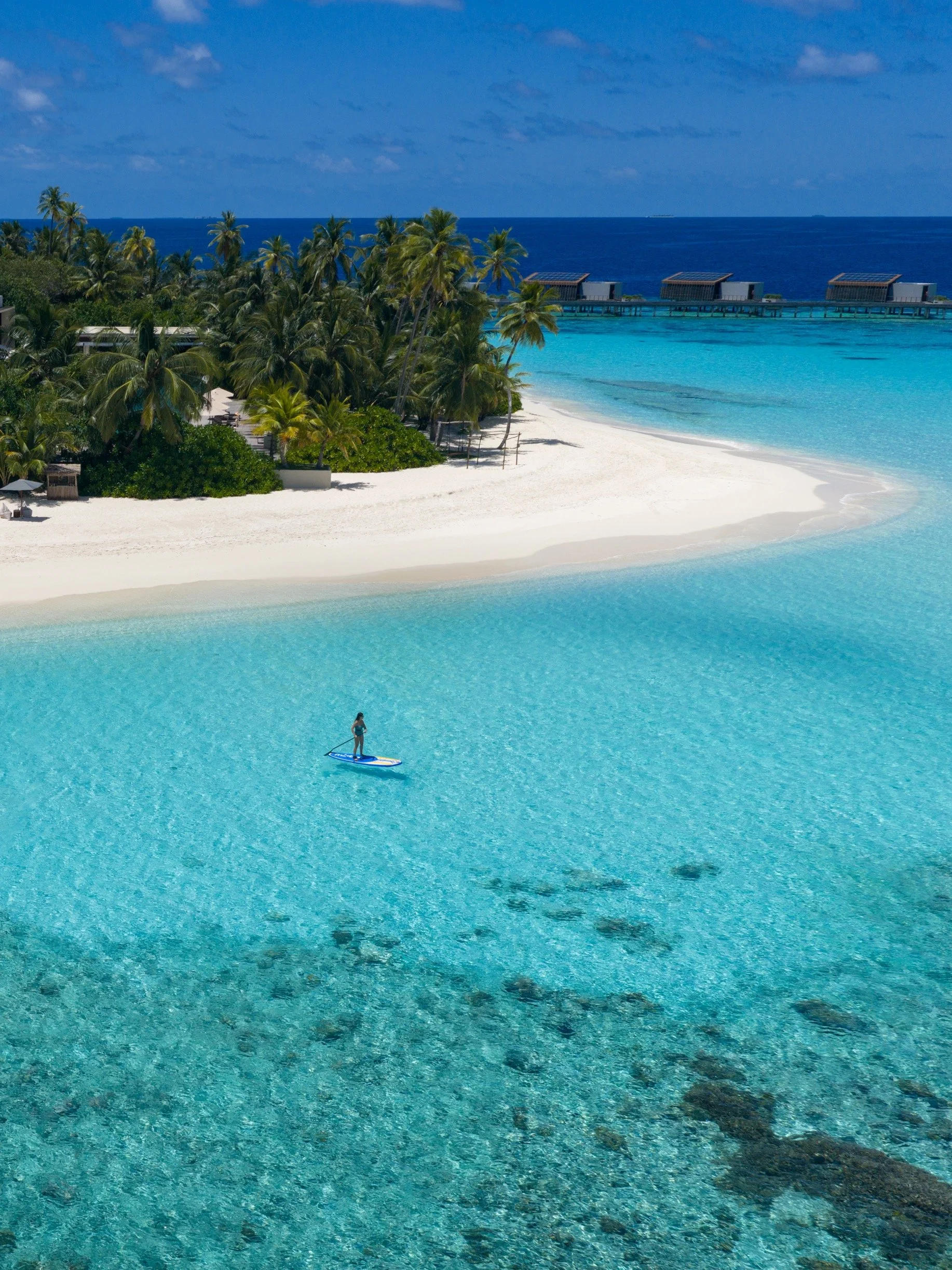 Person paddleboarding on clear turquoise water near a white sandy beach with palm trees and overwater bungalows in the background.