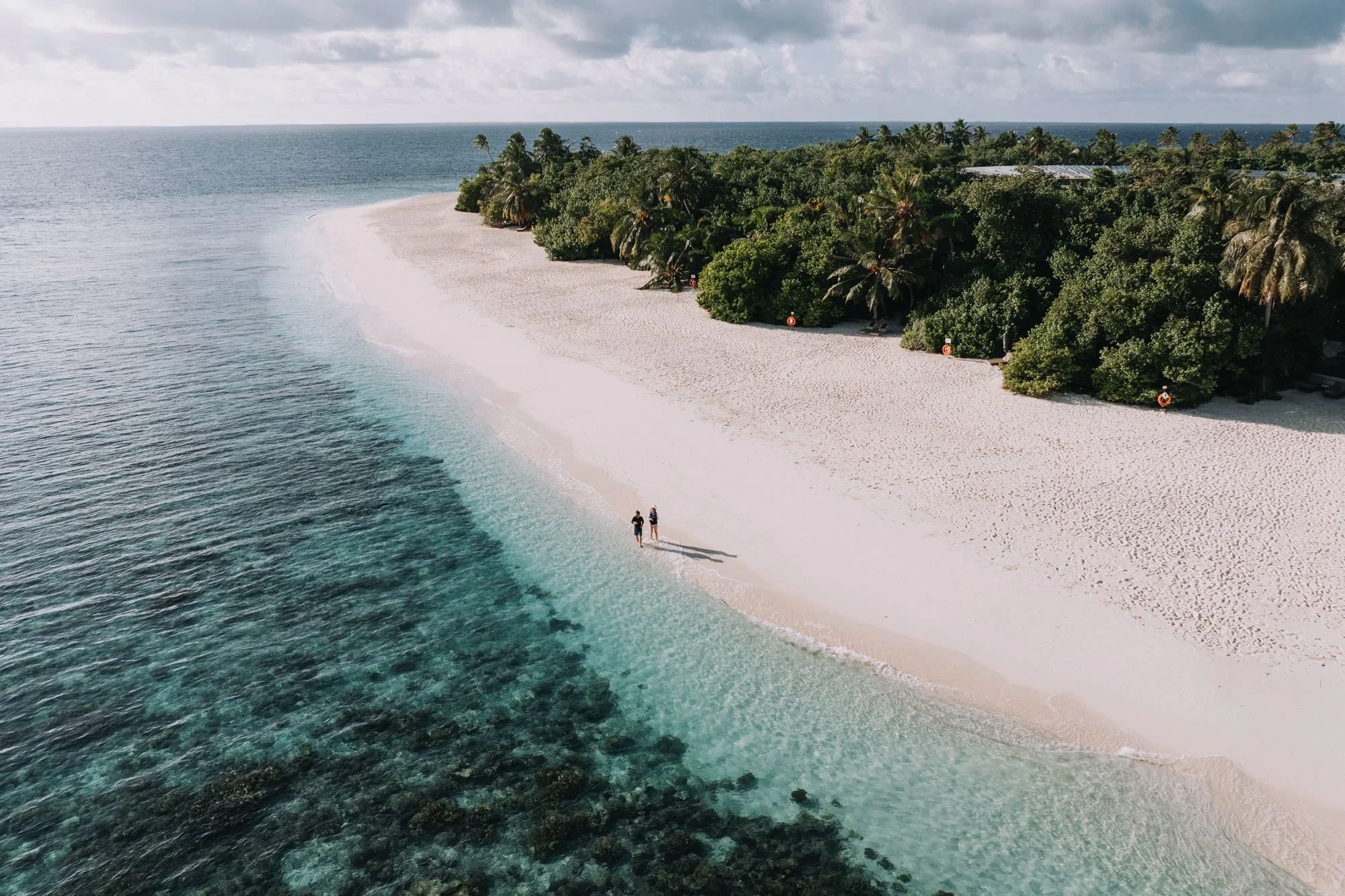 Aerial view of a tropical beach with white sand, clear ocean water, and lush green trees with wind turbines in the background. Two people are walking along the shoreline.