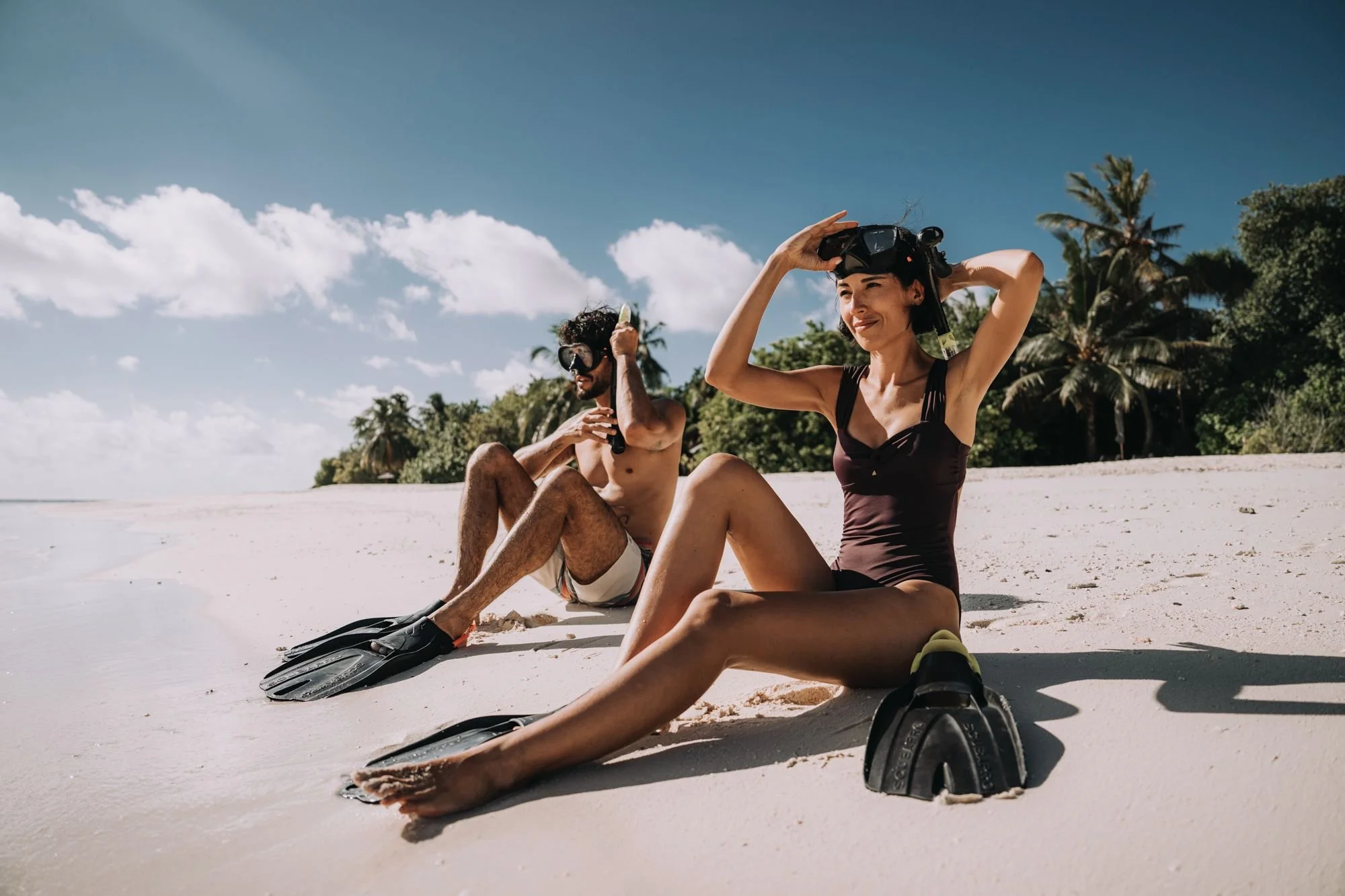 A woman and a man sitting on a sandy beach putting on snorkeling gear, with the woman smiling and the man adjusting his mask, surrounded by palm trees and ocean under a blue sky with clouds.